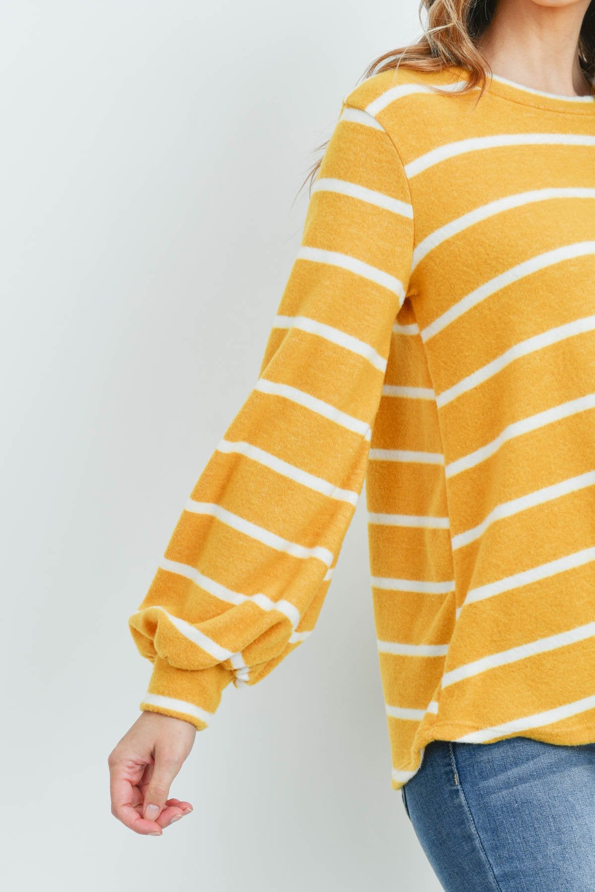 A woman in a Riah Fashion Striped Long Sleeved Round Neck Swing Top and jeans stands against a pink backdrop, wearing a long necklace and touching her hair.