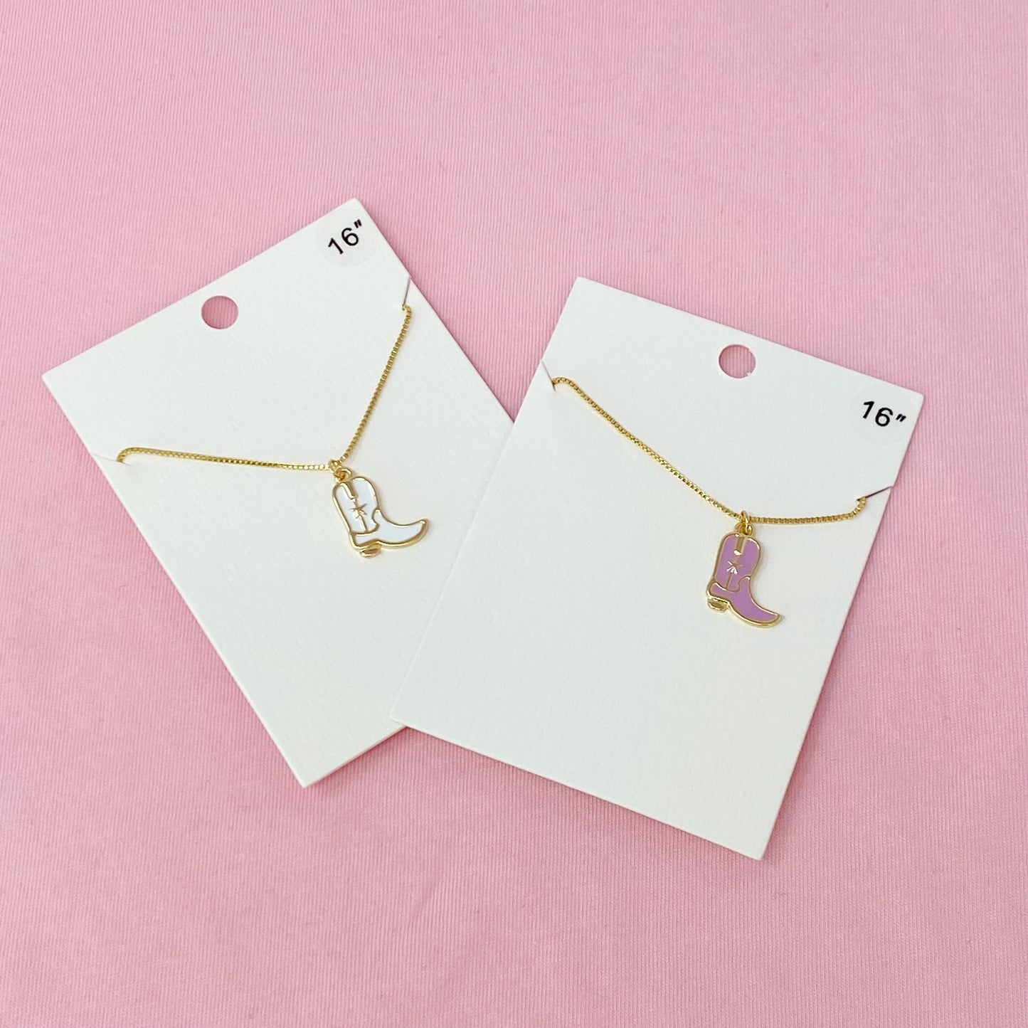 Two Cow Girl Boot Pendant Necklaces by Ellison + Young, one with a white boot and the other with a pink boot, are displayed on a marble surface.