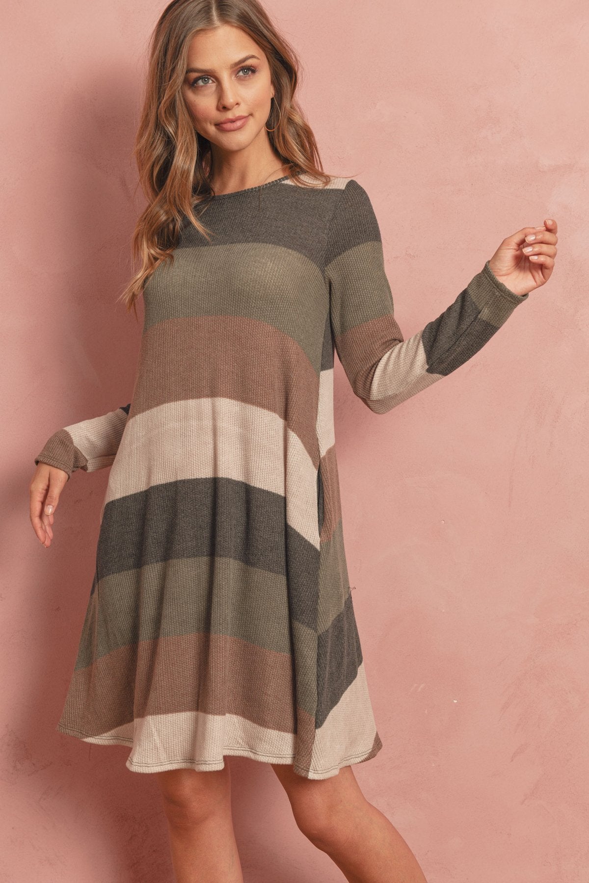 A woman poses in the Riah Fashion Long Sleeved Rib Stripe Pocket Dress in Charcoal/Mocha against a pink textured background.
