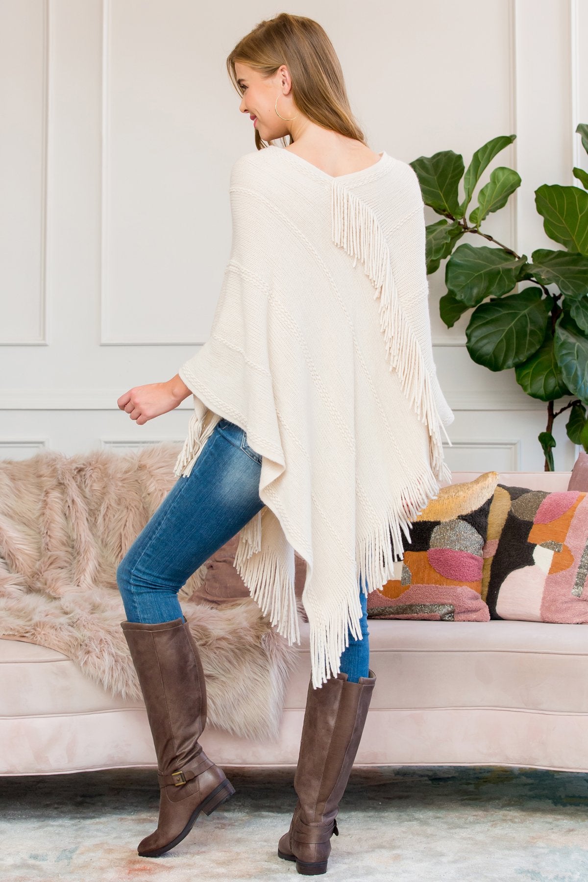 A person stands wearing a Riah Fashion Asymmetric Fringe Poncho in one color, paired with jeans and brown boots, smiling while holding the poncho. In the background are a plant and a sofa with cushions.