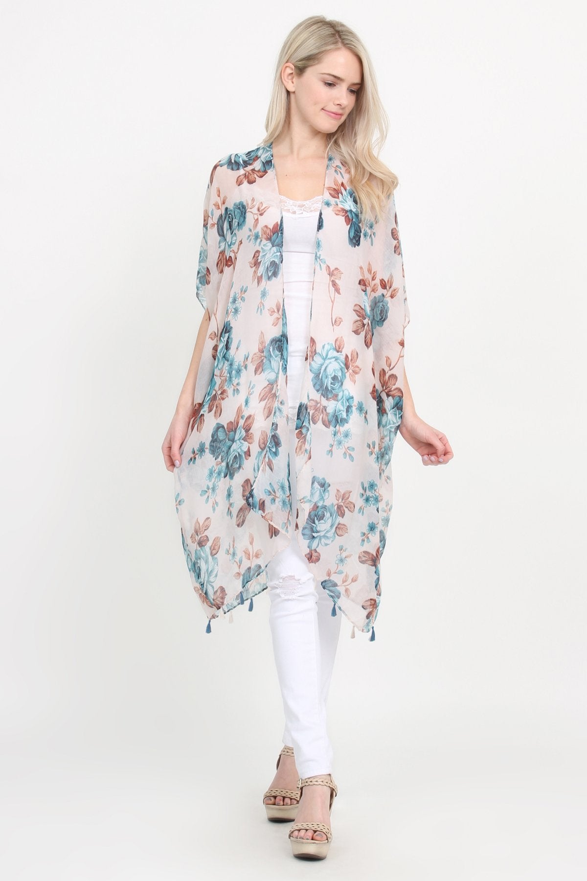A woman stands against a plain background, wearing a Riah Fashion beige blue open front knee-length floral cardigan over a white outfit.