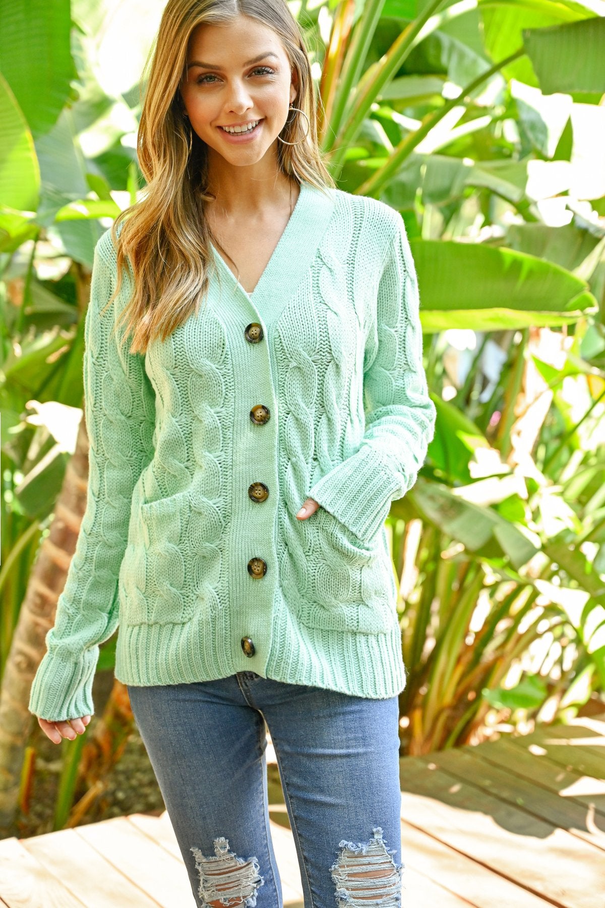 A person wearing an ash copper cable sweater with pockets from Riah Fashion stands in front of tall green plants and a wooden fence.