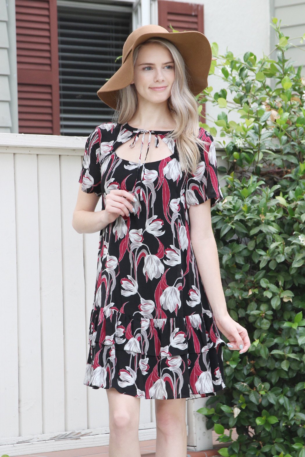 A woman in a Riah Fashion Short Sleeve Floral Ruffle Dress and a brown hat stands near a wooden fence and greenery.