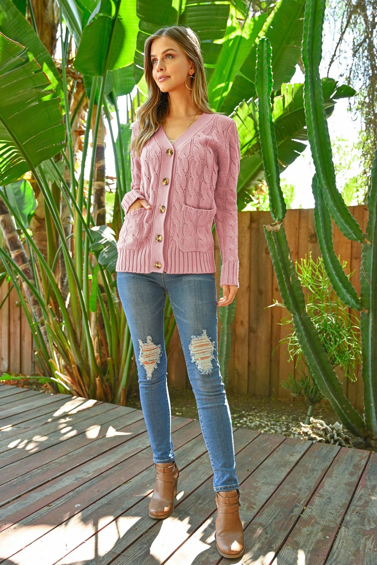 A person wearing an ash copper cable sweater with pockets from Riah Fashion stands in front of tall green plants and a wooden fence.