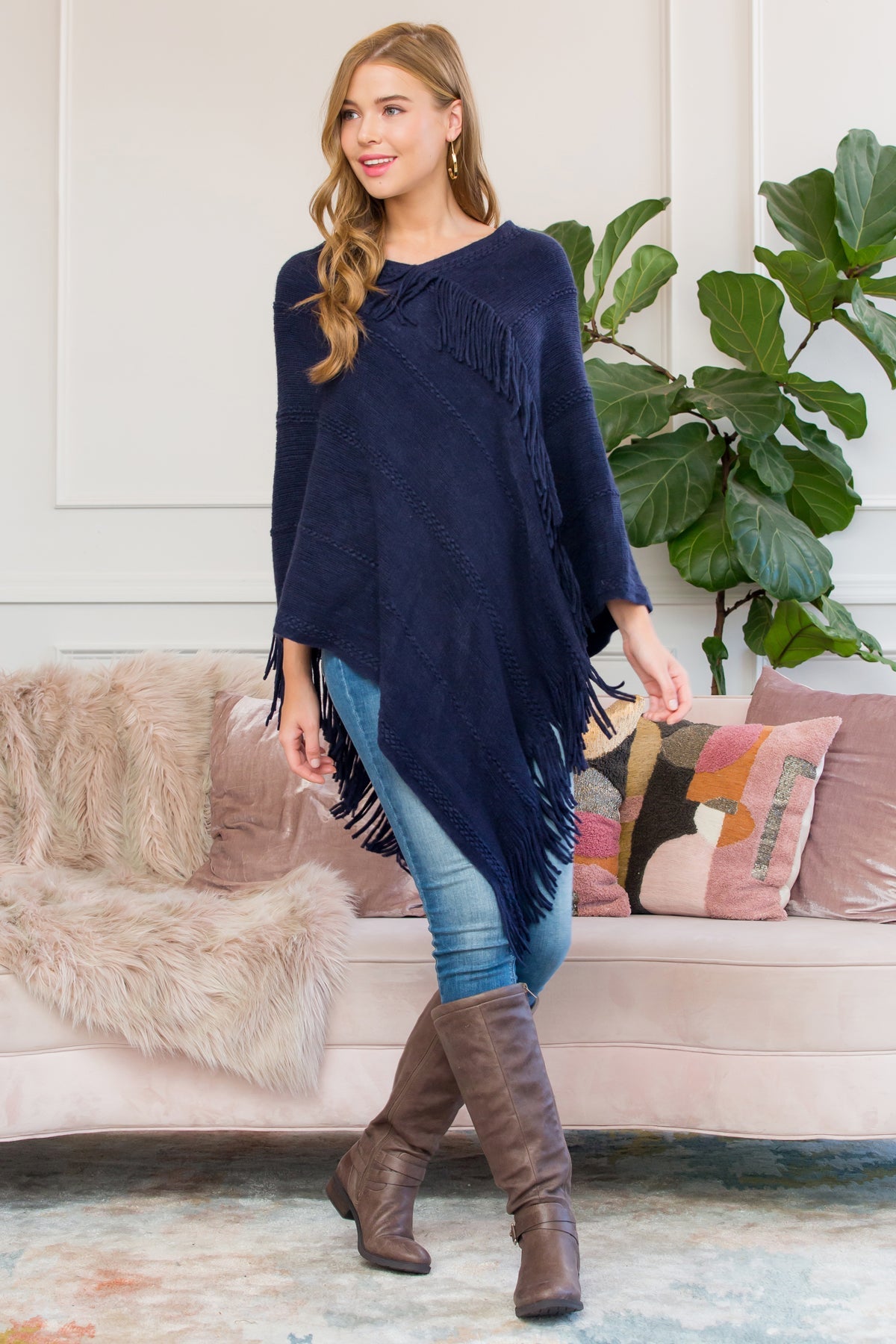 A person stands wearing a Riah Fashion Asymmetric Fringe Poncho in one color, paired with jeans and brown boots, smiling while holding the poncho. In the background are a plant and a sofa with cushions.