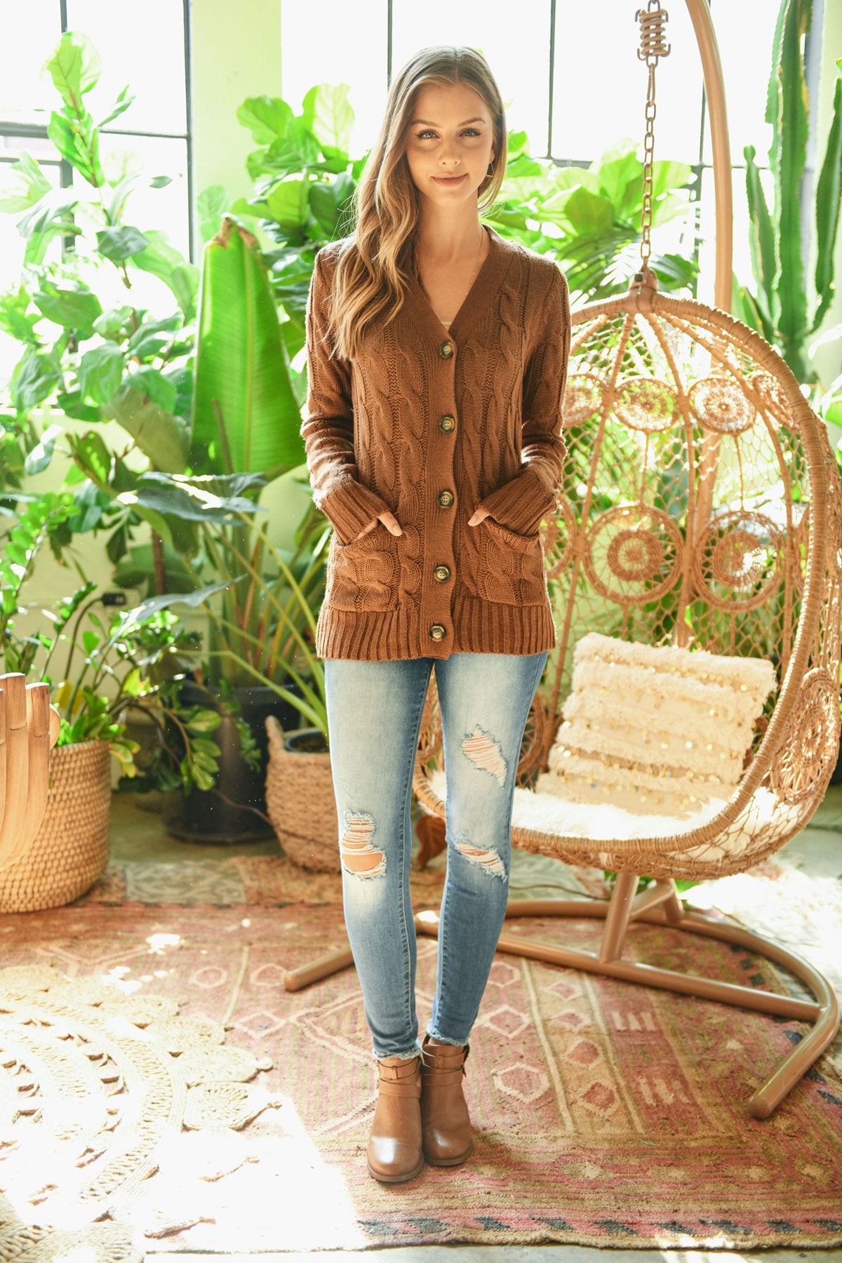 A person wearing an ash copper cable sweater with pockets from Riah Fashion stands in front of tall green plants and a wooden fence.