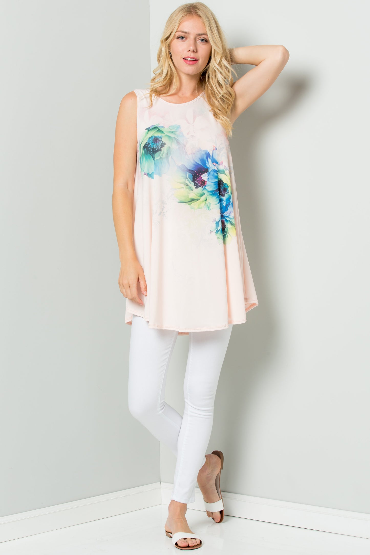 A person styled in a Sleeveless Tunic Dress With Floral Print Sublimation by Riah Fashion, paired with white pants, stands against a plain backdrop.