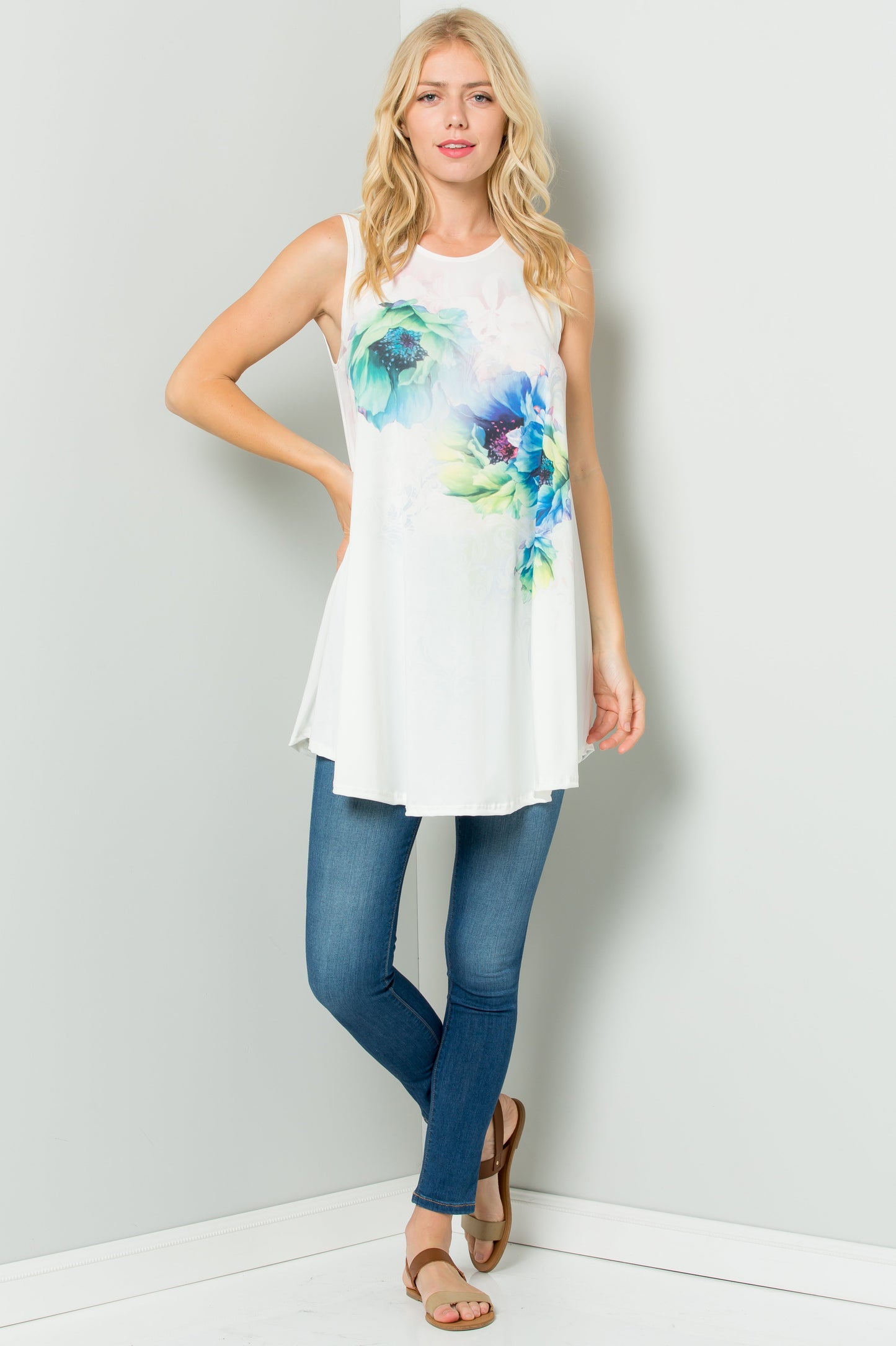 A person styled in a Sleeveless Tunic Dress With Floral Print Sublimation by Riah Fashion, paired with white pants, stands against a plain backdrop.