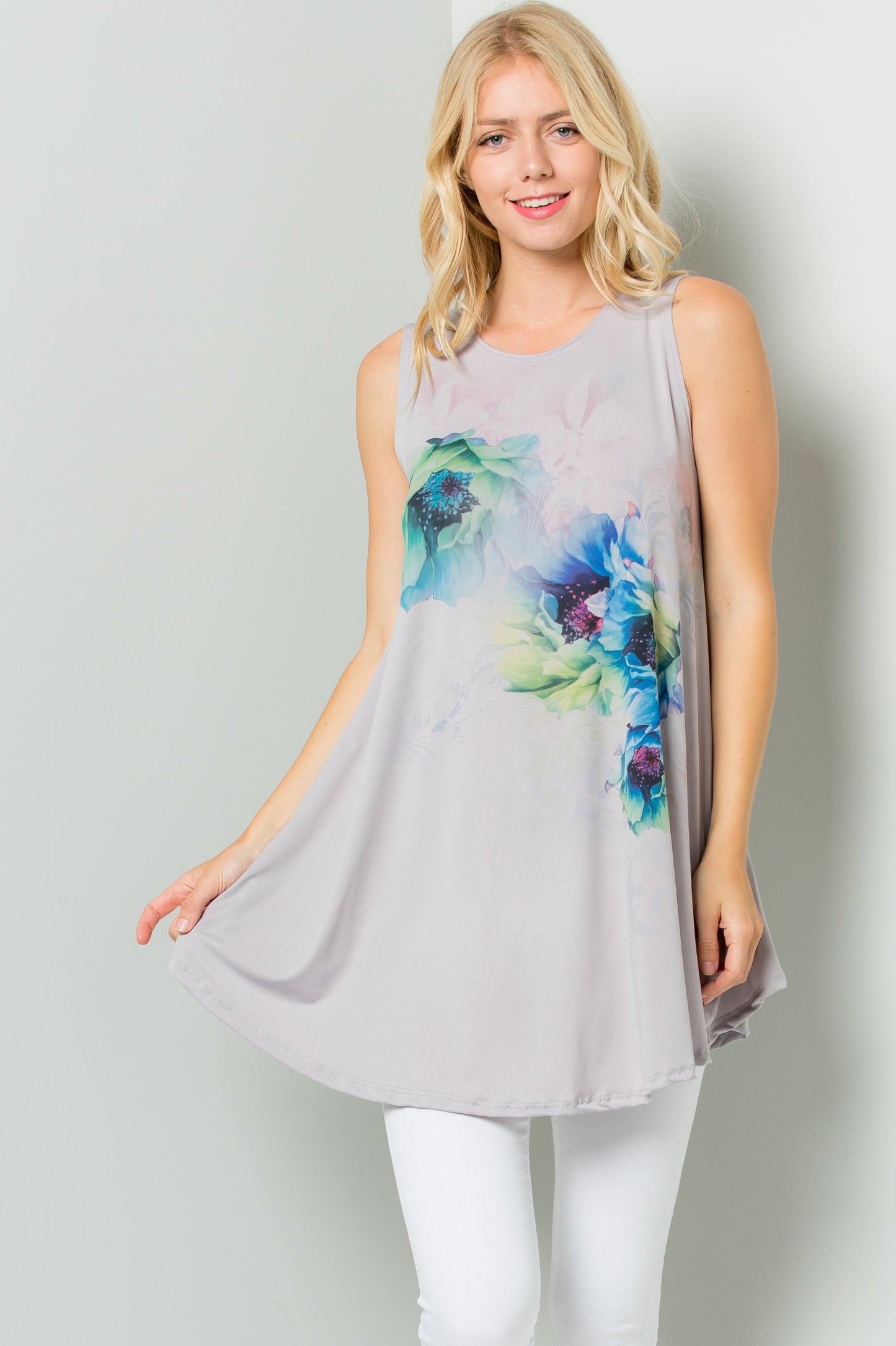A person styled in a Sleeveless Tunic Dress With Floral Print Sublimation by Riah Fashion, paired with white pants, stands against a plain backdrop.
