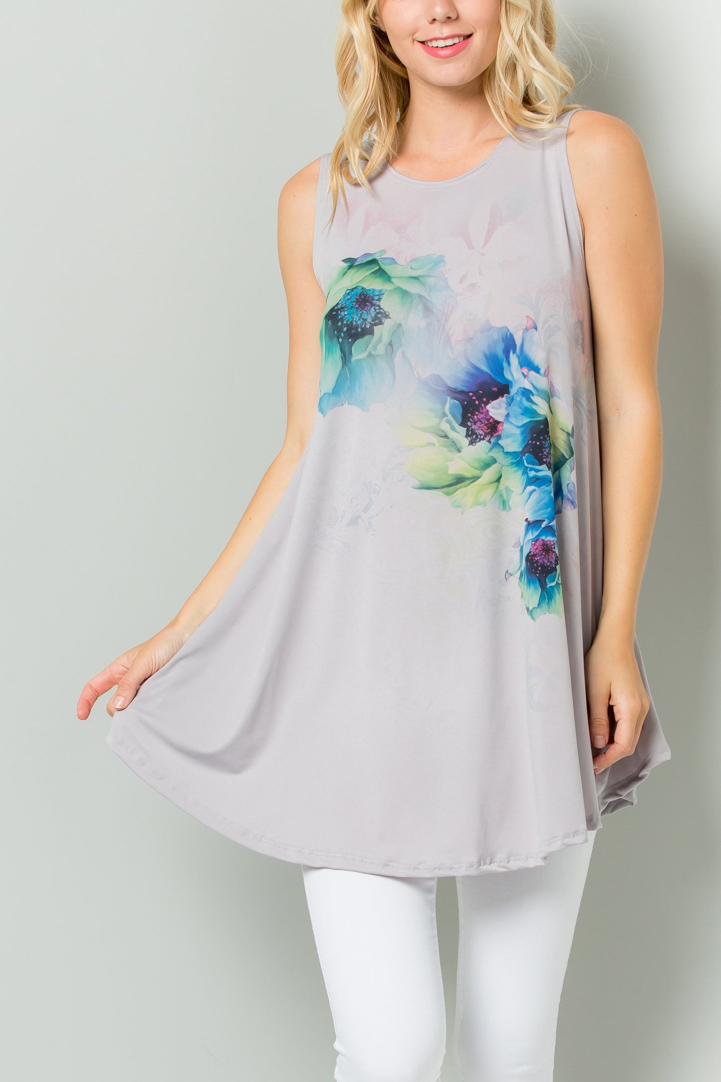 A person styled in a Sleeveless Tunic Dress With Floral Print Sublimation by Riah Fashion, paired with white pants, stands against a plain backdrop.
