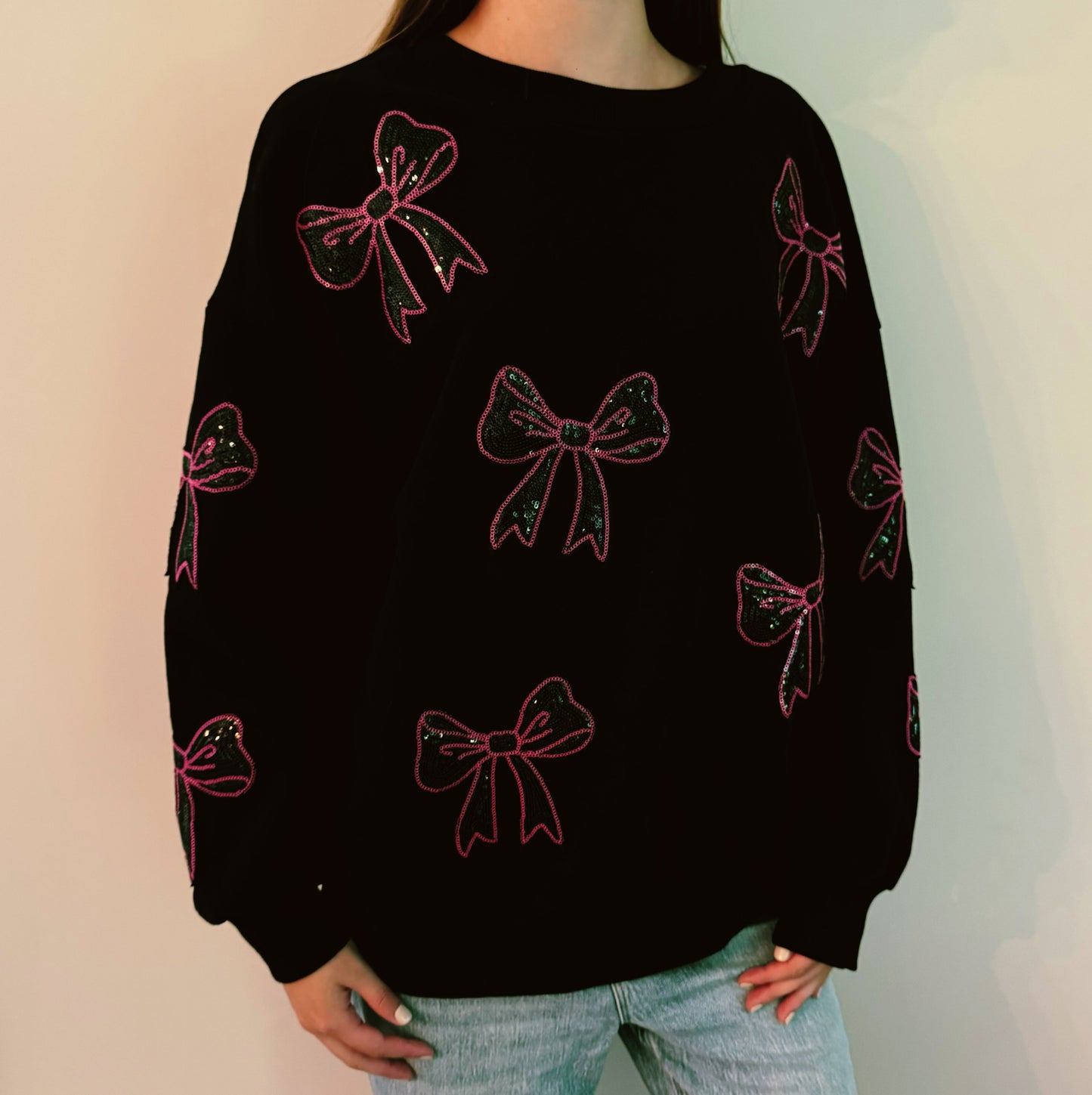 A person with long hair sits on a couch wearing an Ellison + Young Sparkly Bow Patch Sweatshirt paired with jeans, smiling and resting one hand near their face.
