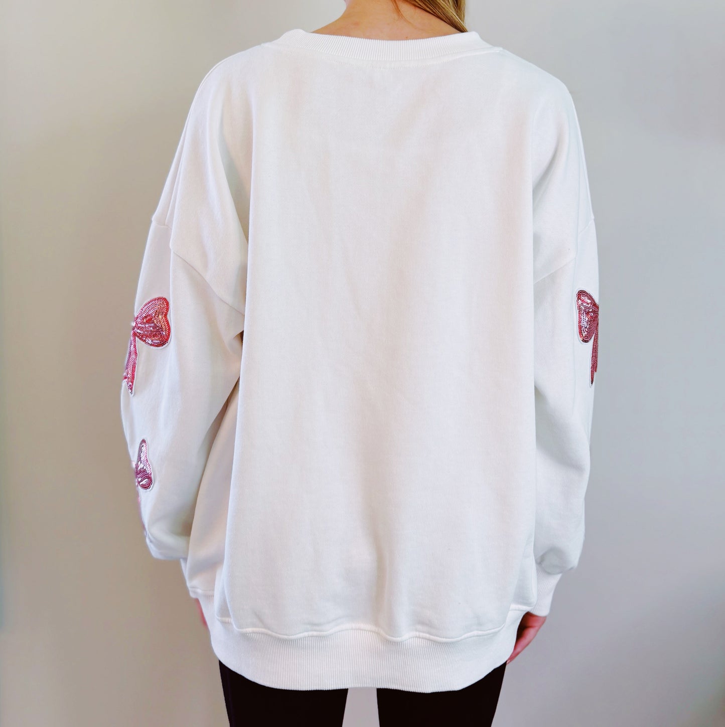 A person with long hair sits on a couch wearing an Ellison + Young Sparkly Bow Patch Sweatshirt paired with jeans, smiling and resting one hand near their face.