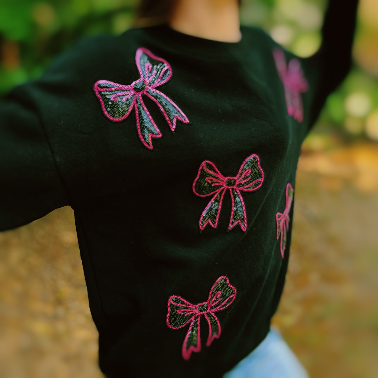 A person with long hair sits on a couch wearing an Ellison + Young Sparkly Bow Patch Sweatshirt paired with jeans, smiling and resting one hand near their face.