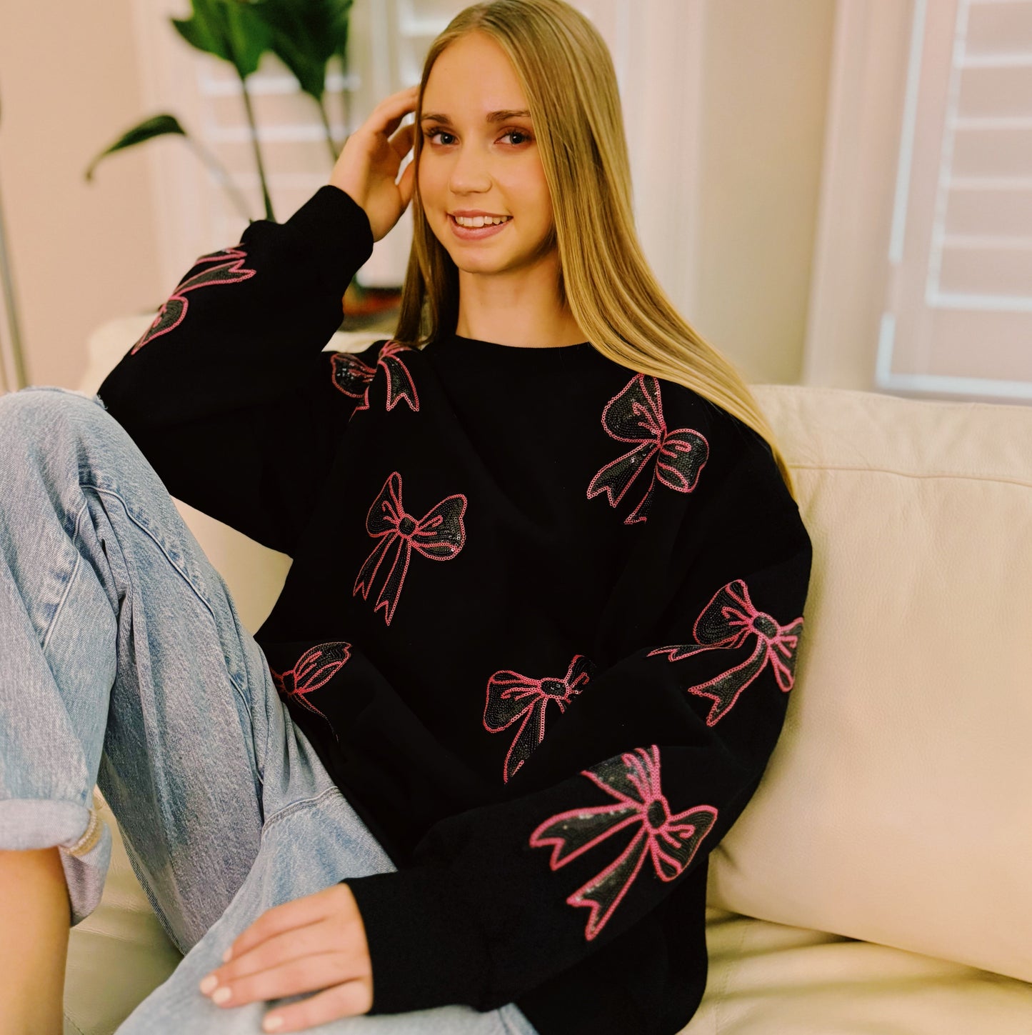 A person with long hair sits on a couch wearing an Ellison + Young Sparkly Bow Patch Sweatshirt paired with jeans, smiling and resting one hand near their face.