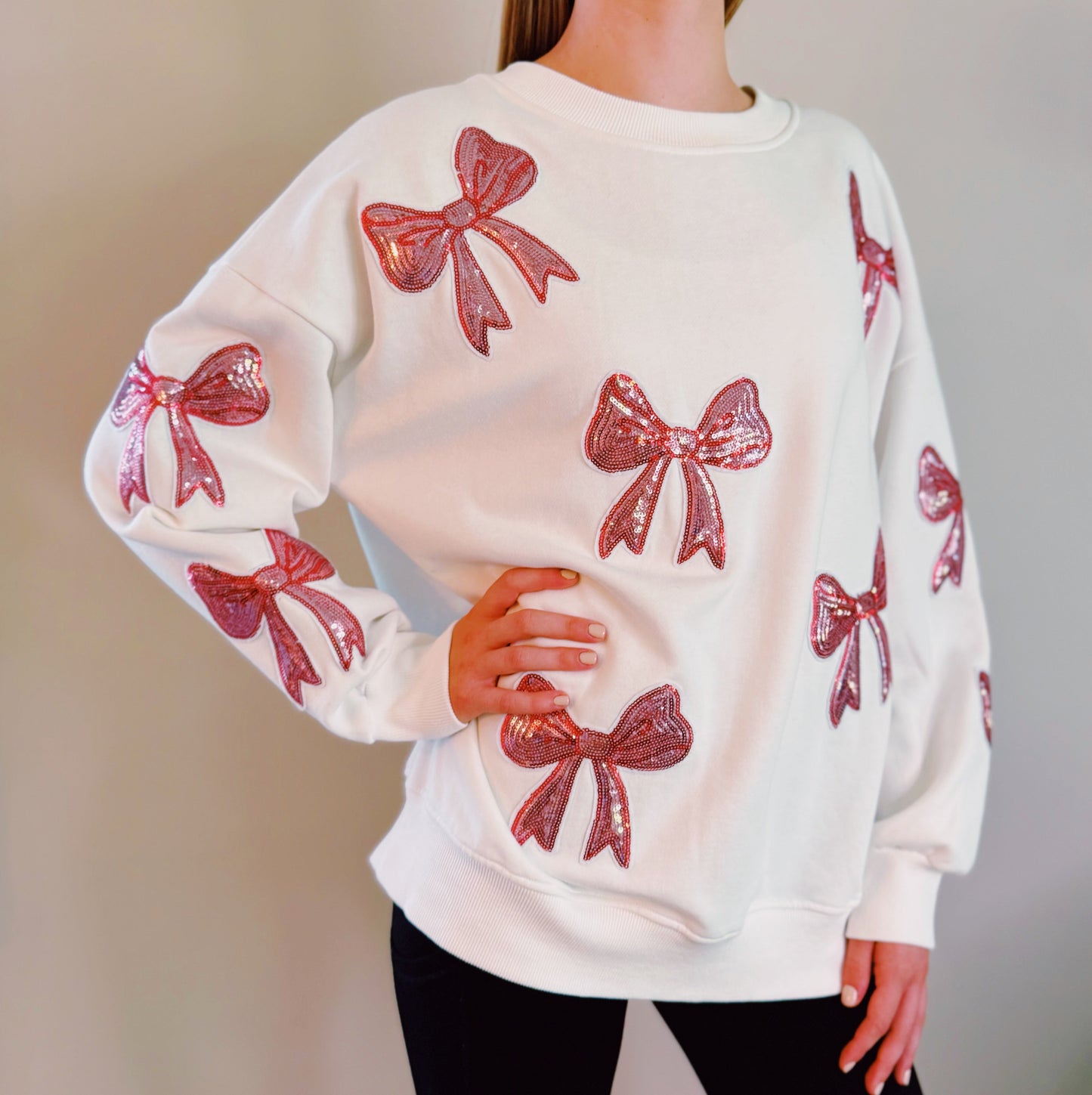 A person with long hair sits on a couch wearing an Ellison + Young Sparkly Bow Patch Sweatshirt paired with jeans, smiling and resting one hand near their face.