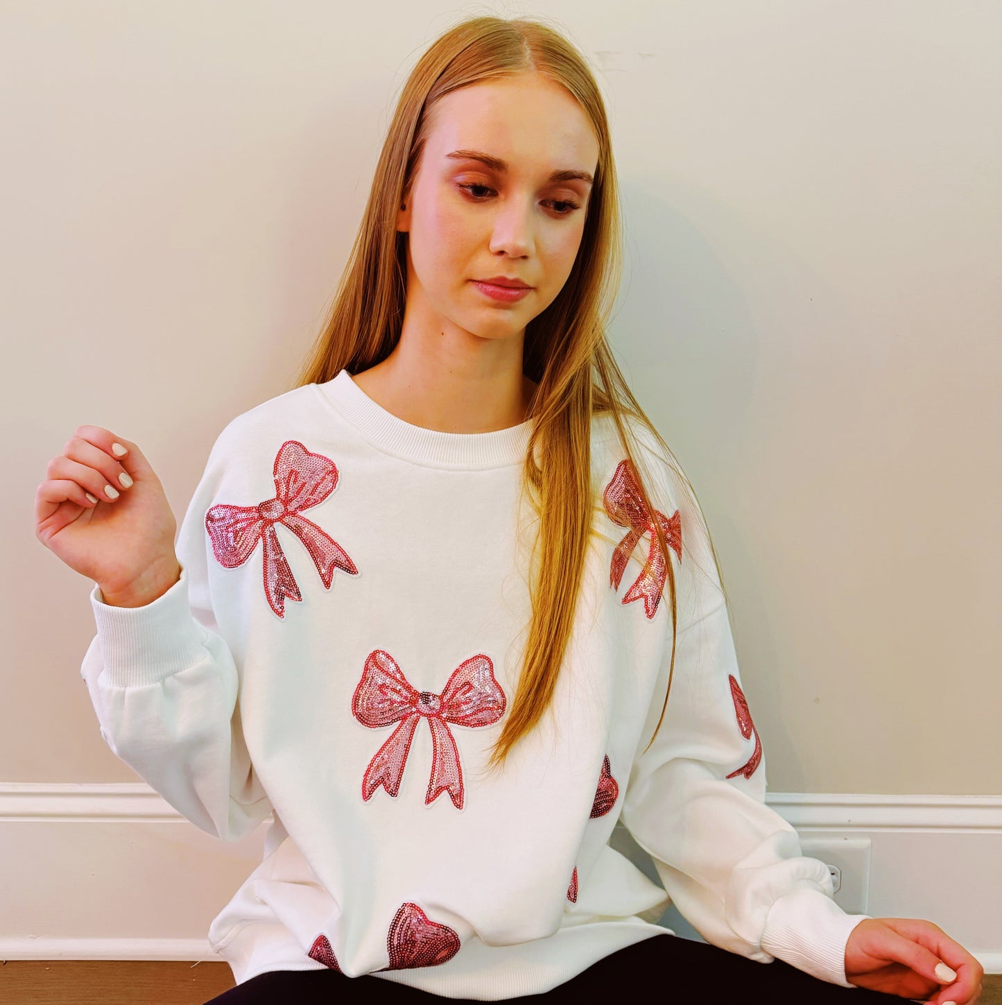 A person with long hair sits on a couch wearing an Ellison + Young Sparkly Bow Patch Sweatshirt paired with jeans, smiling and resting one hand near their face.