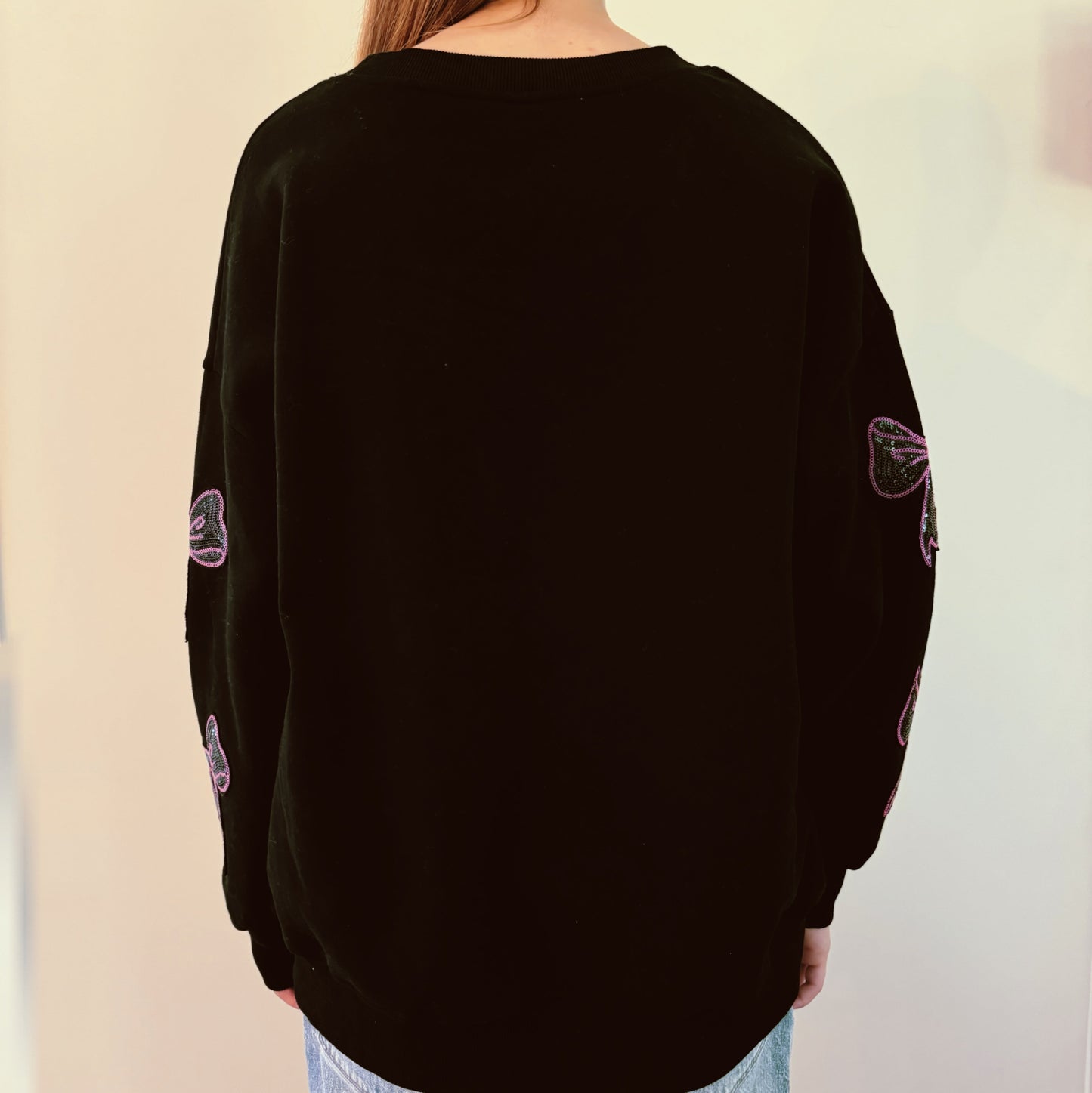 A person with long hair sits on a couch wearing an Ellison + Young Sparkly Bow Patch Sweatshirt paired with jeans, smiling and resting one hand near their face.