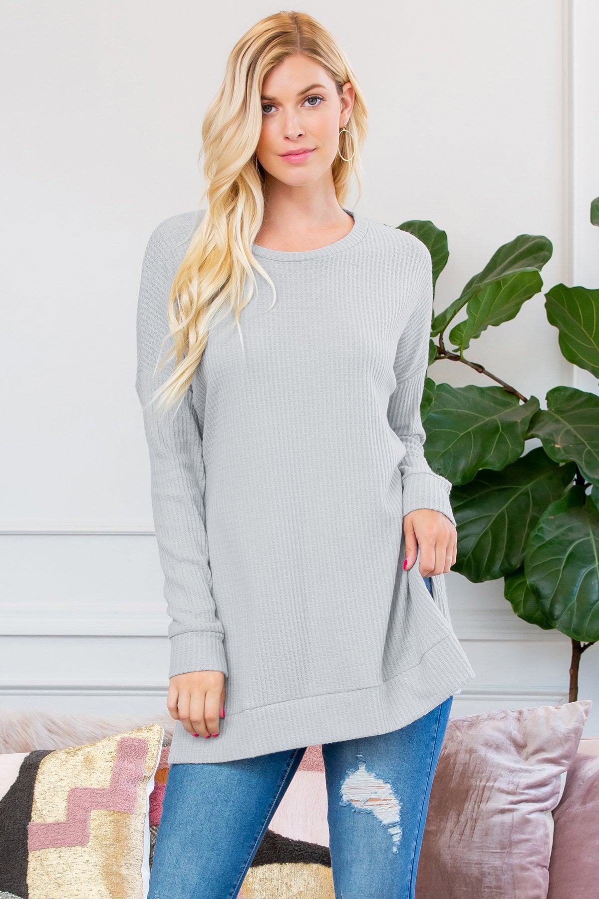 A woman stands on a wooden porch wearing Riah Fashions Brushed Thermal Waffle Sweater and jeans, accessorized with a necklace. Greenery and a wooden fence are visible in the background.