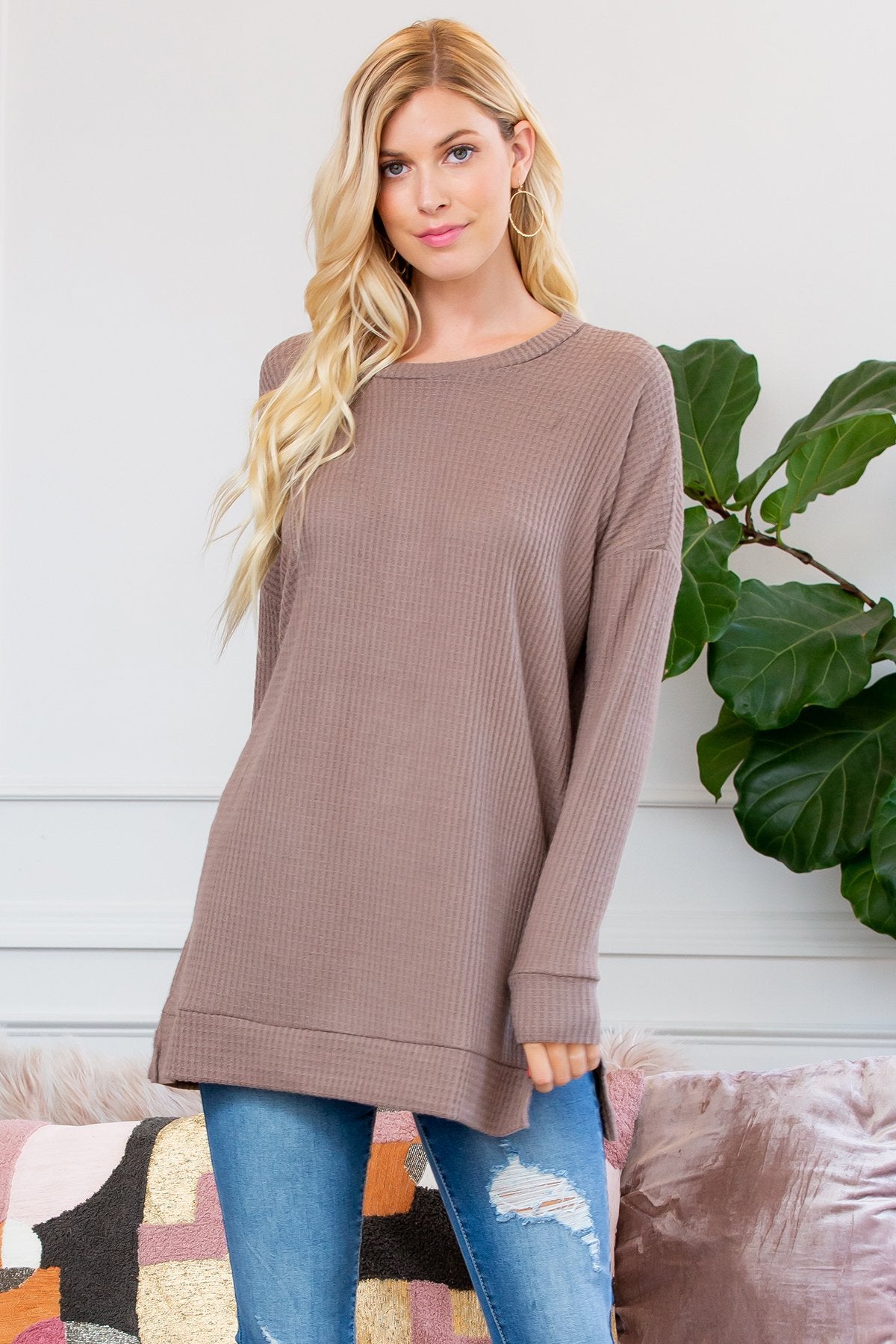 A woman stands on a wooden porch wearing Riah Fashions Brushed Thermal Waffle Sweater and jeans, accessorized with a necklace. Greenery and a wooden fence are visible in the background.
