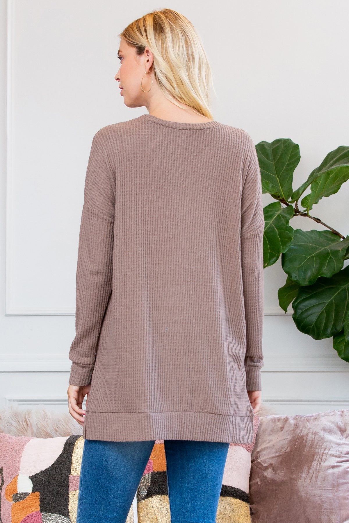 A woman stands on a wooden porch wearing Riah Fashions Brushed Thermal Waffle Sweater and jeans, accessorized with a necklace. Greenery and a wooden fence are visible in the background.