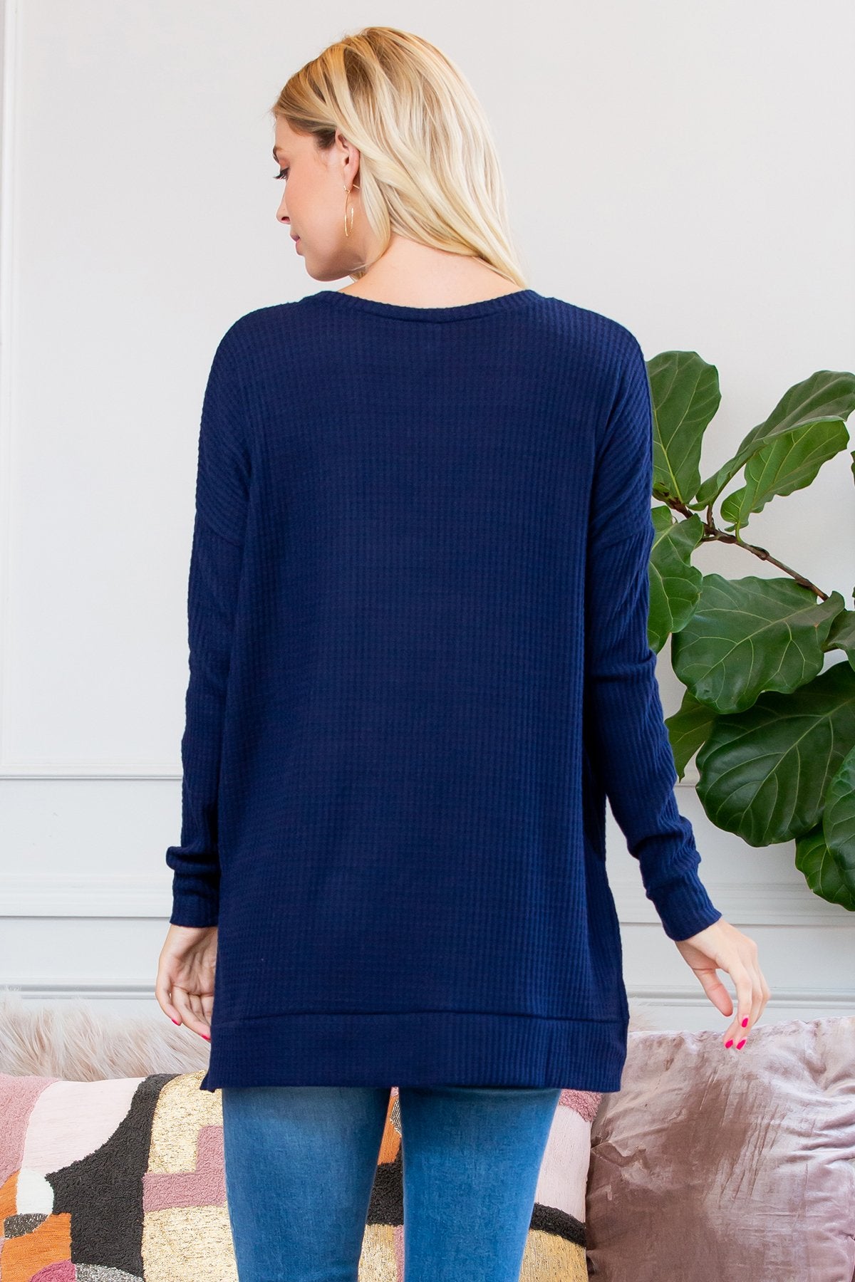 A woman stands on a wooden porch wearing Riah Fashions Brushed Thermal Waffle Sweater and jeans, accessorized with a necklace. Greenery and a wooden fence are visible in the background.