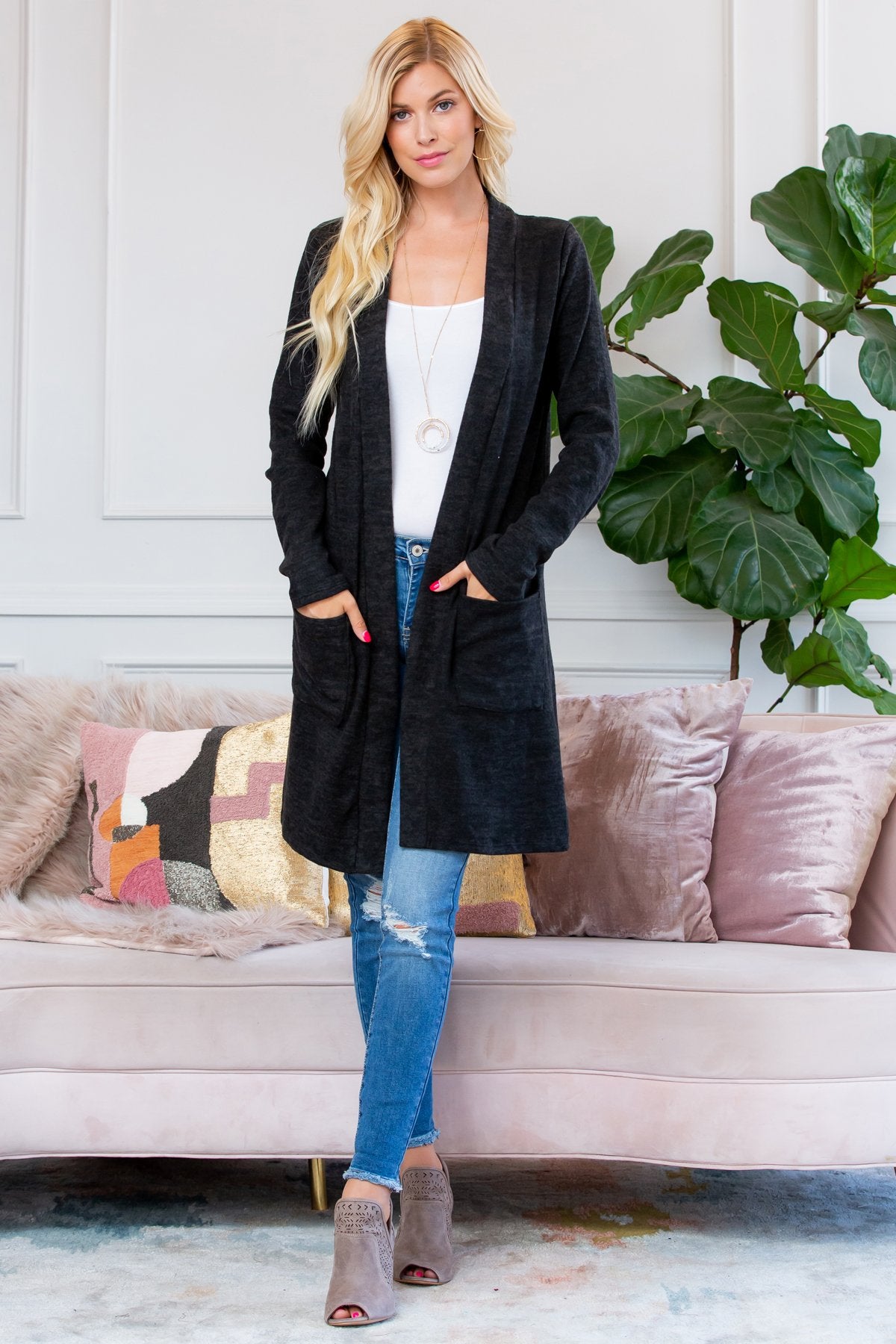 A person stands indoors near a plant, wearing Riah Fashions Brushed Sweater Pocket Cardigan in black over a white top and jeans.