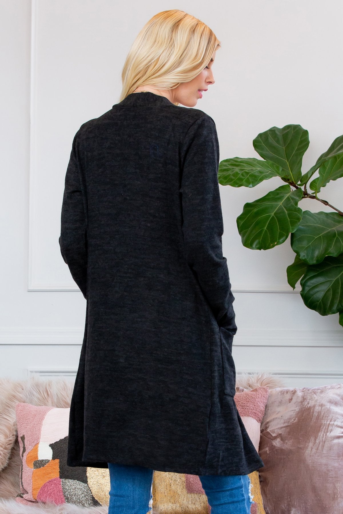 A person stands indoors near a plant, wearing Riah Fashions Brushed Sweater Pocket Cardigan in black over a white top and jeans.
