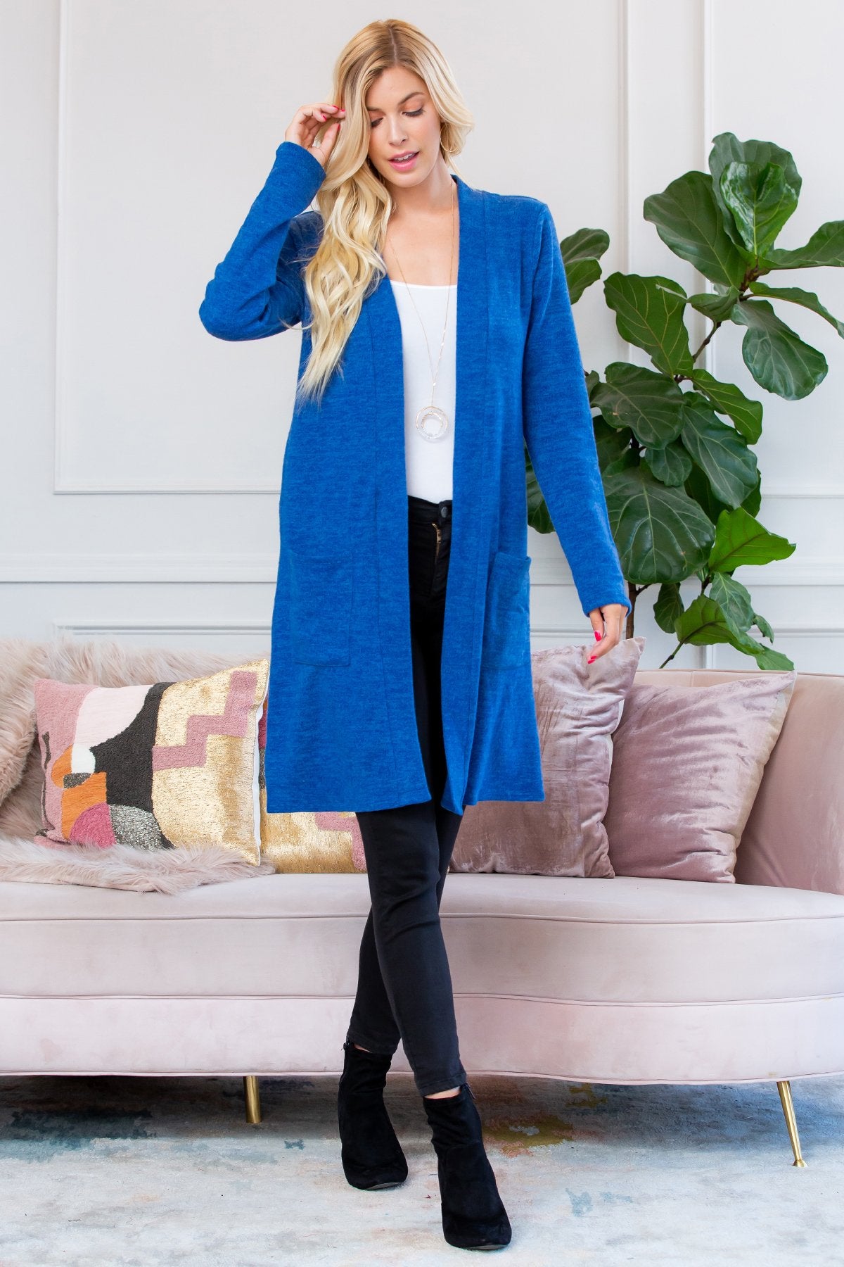 A person stands indoors near a plant, wearing Riah Fashions Brushed Sweater Pocket Cardigan in black over a white top and jeans.