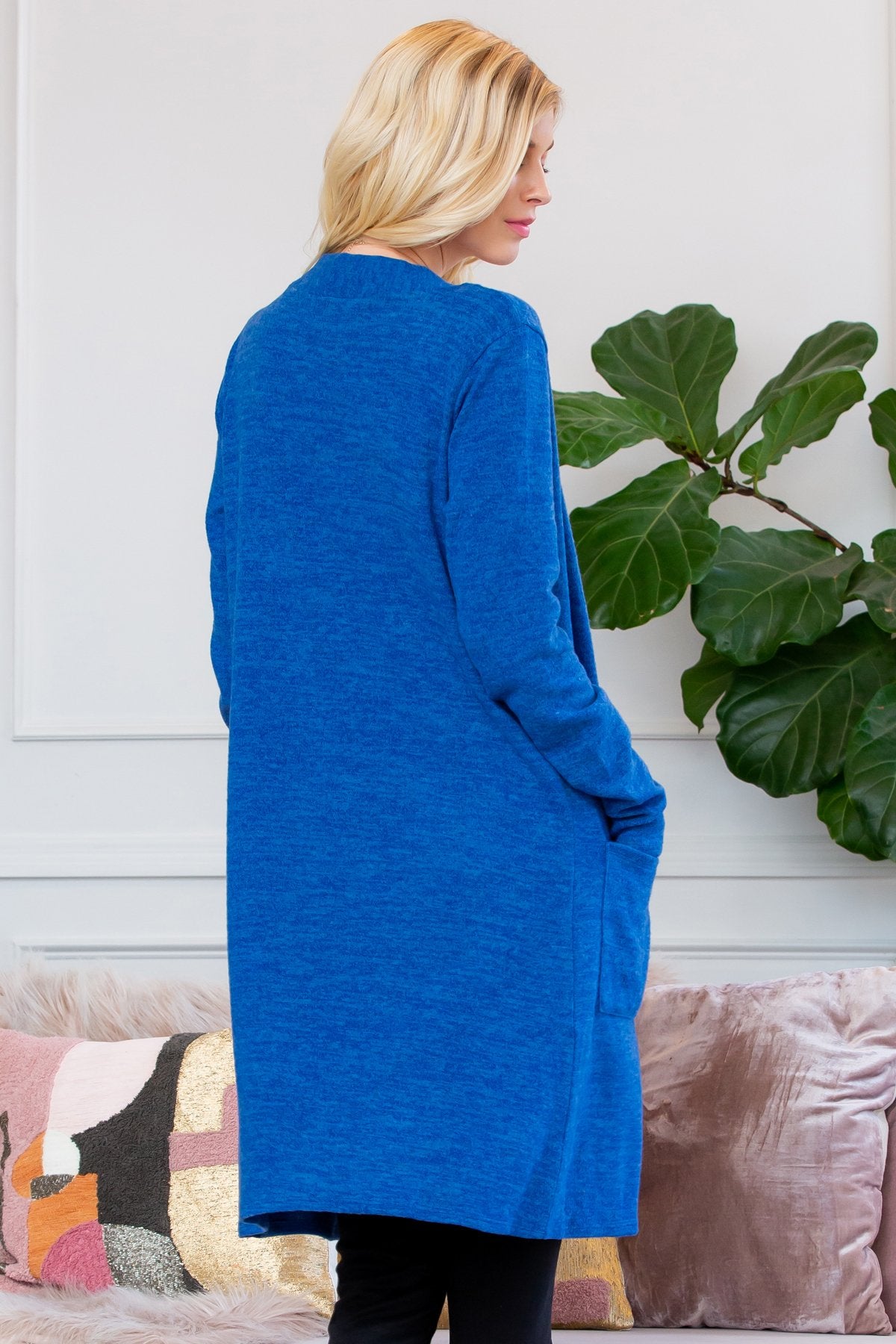 A person stands indoors near a plant, wearing Riah Fashions Brushed Sweater Pocket Cardigan in black over a white top and jeans.