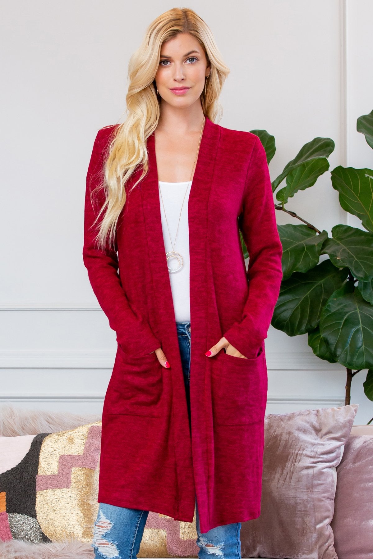 A person stands indoors near a plant, wearing Riah Fashions Brushed Sweater Pocket Cardigan in black over a white top and jeans.