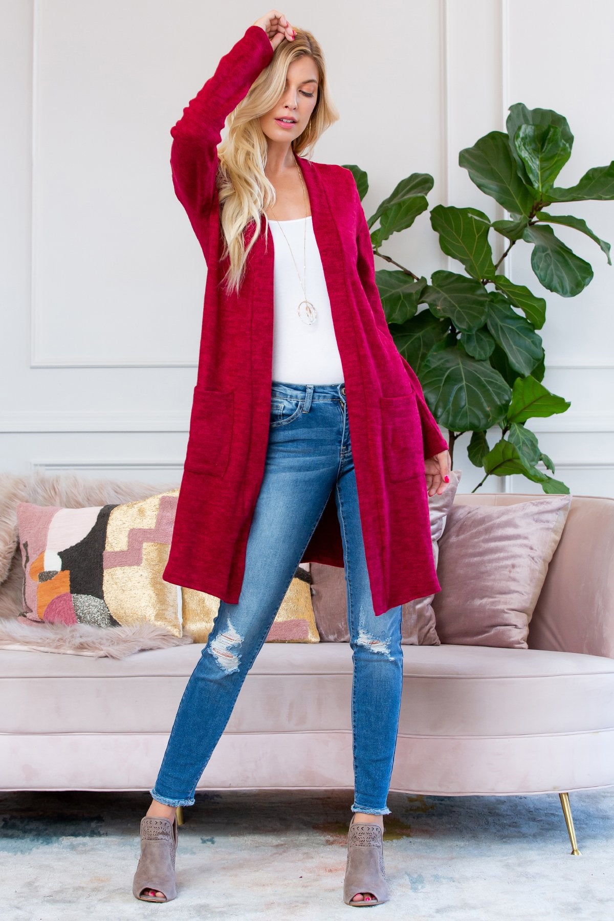 A person stands indoors near a plant, wearing Riah Fashions Brushed Sweater Pocket Cardigan in black over a white top and jeans.