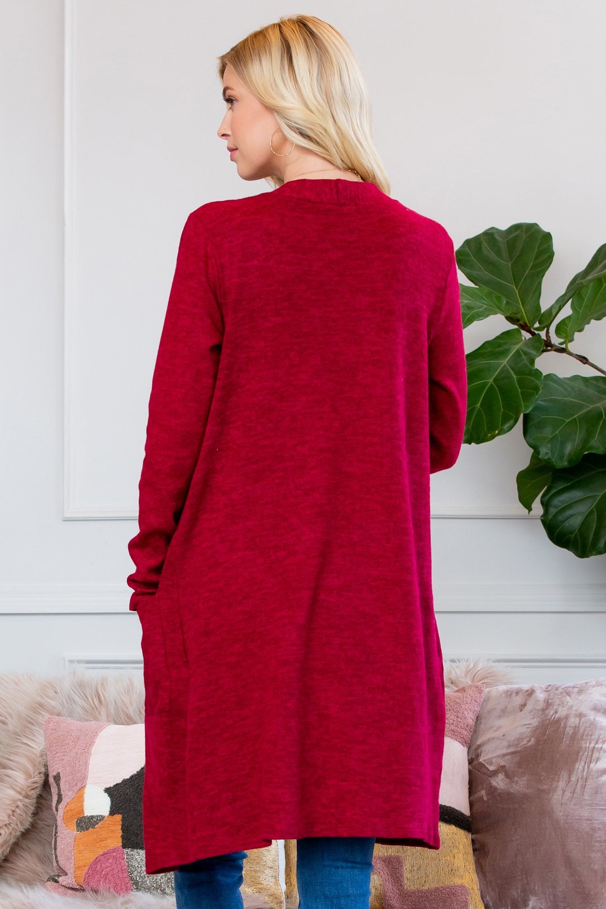 A person stands indoors near a plant, wearing Riah Fashions Brushed Sweater Pocket Cardigan in black over a white top and jeans.