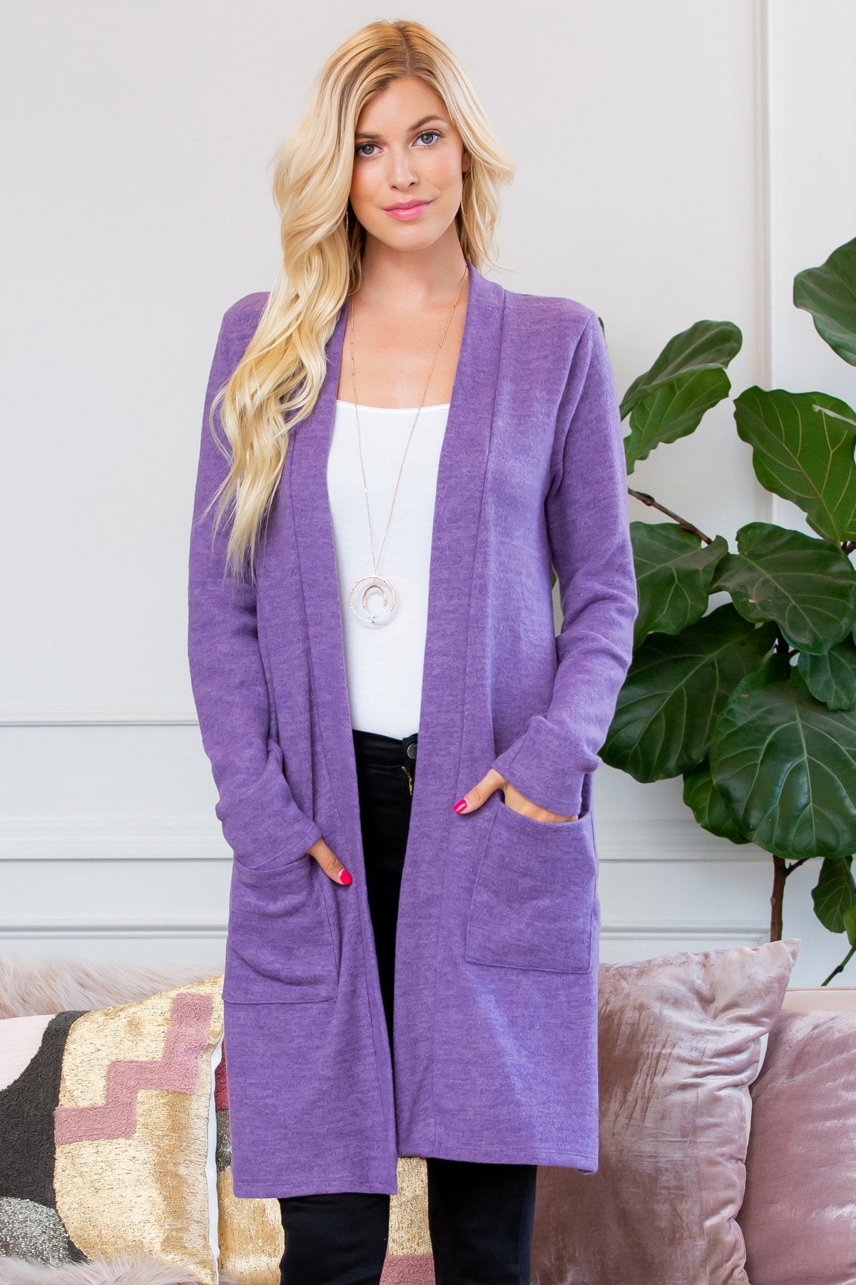 A person stands indoors near a plant, wearing Riah Fashions Brushed Sweater Pocket Cardigan in black over a white top and jeans.