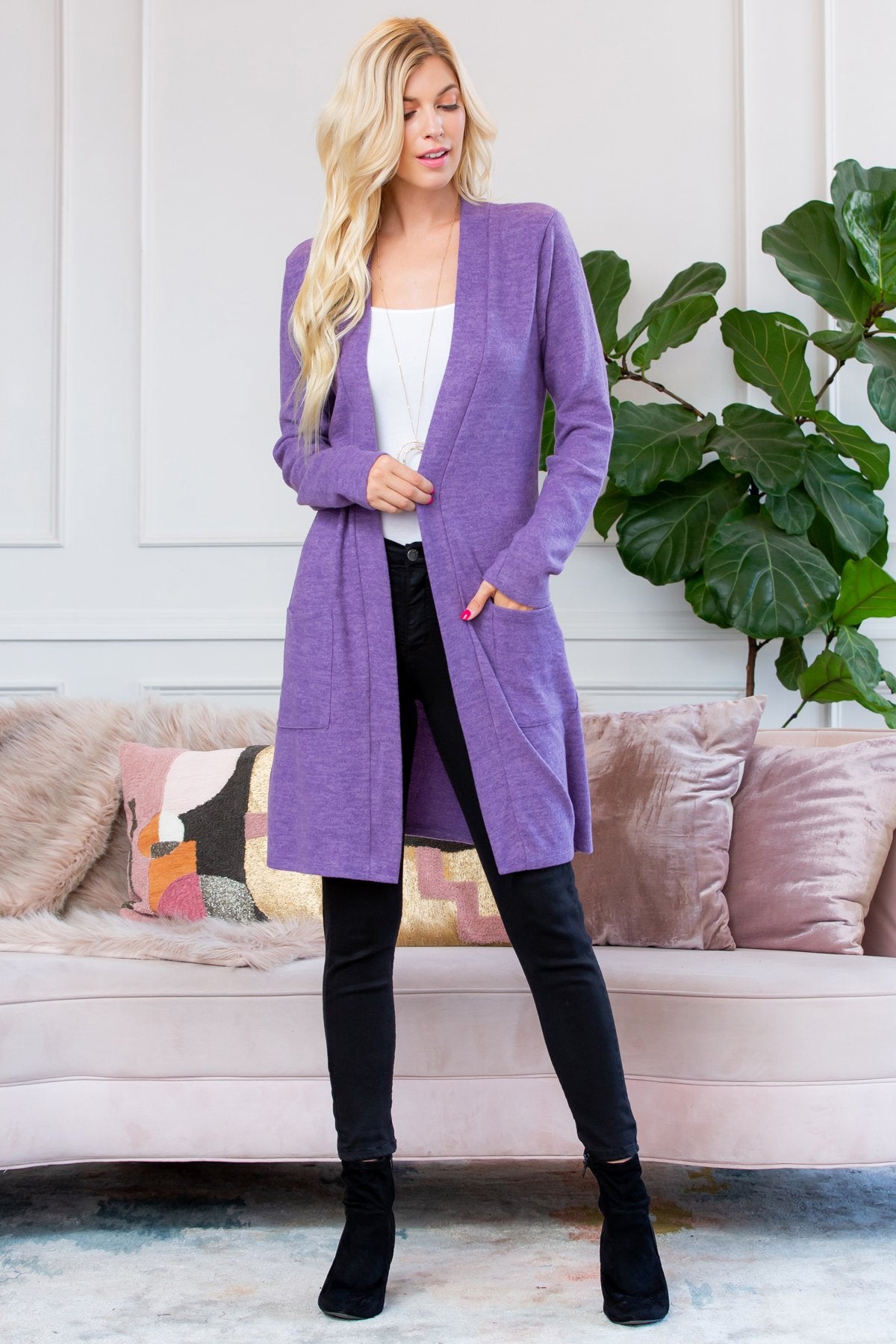 A person stands indoors near a plant, wearing Riah Fashions Brushed Sweater Pocket Cardigan in black over a white top and jeans.