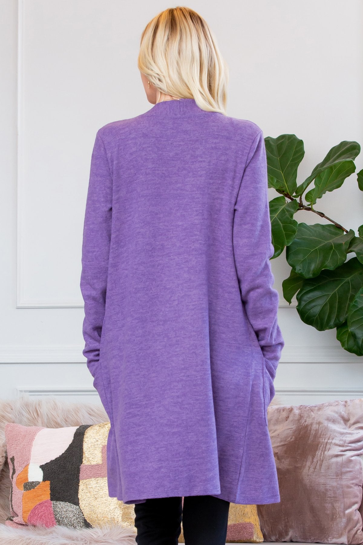 A person stands indoors near a plant, wearing Riah Fashions Brushed Sweater Pocket Cardigan in black over a white top and jeans.