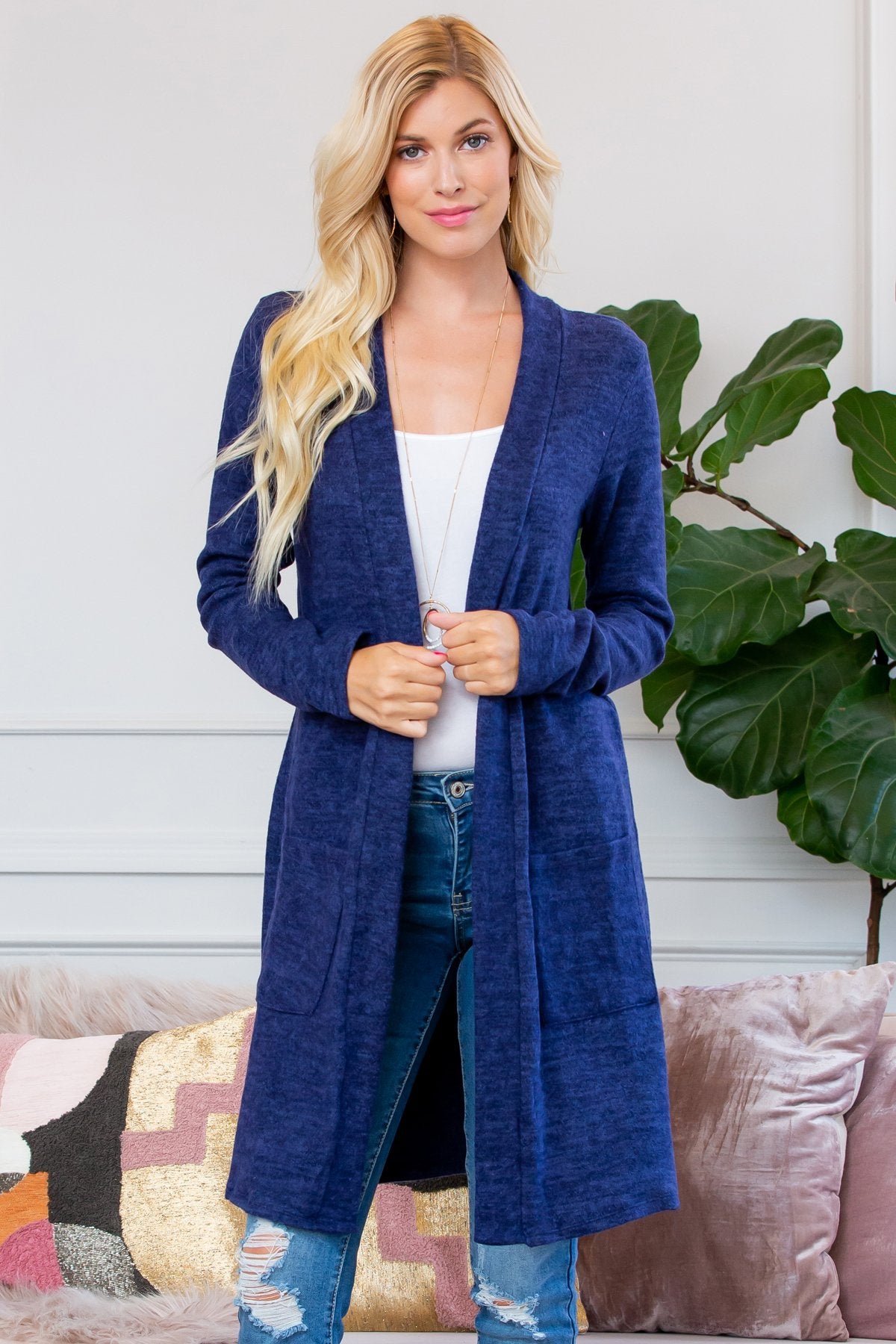 A person stands indoors near a plant, wearing Riah Fashions Brushed Sweater Pocket Cardigan in black over a white top and jeans.