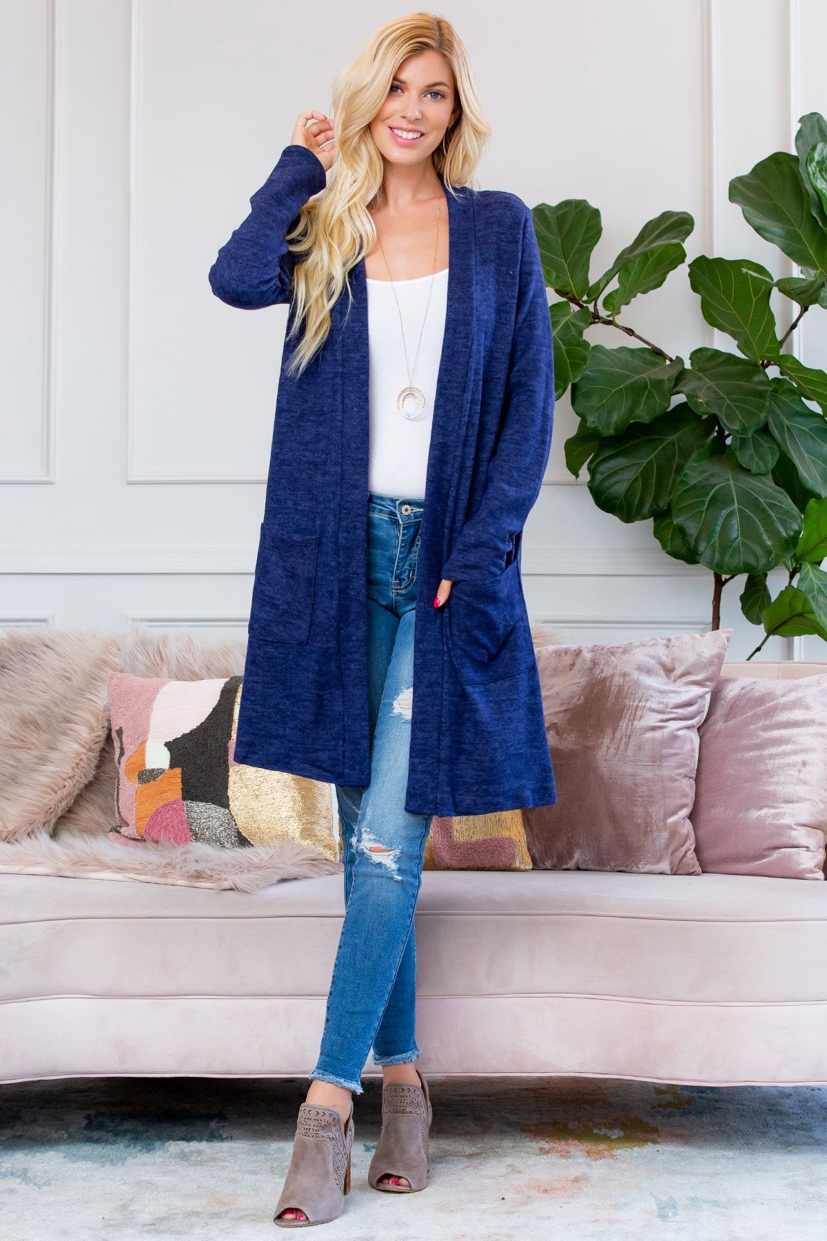 A person stands indoors near a plant, wearing Riah Fashions Brushed Sweater Pocket Cardigan in black over a white top and jeans.