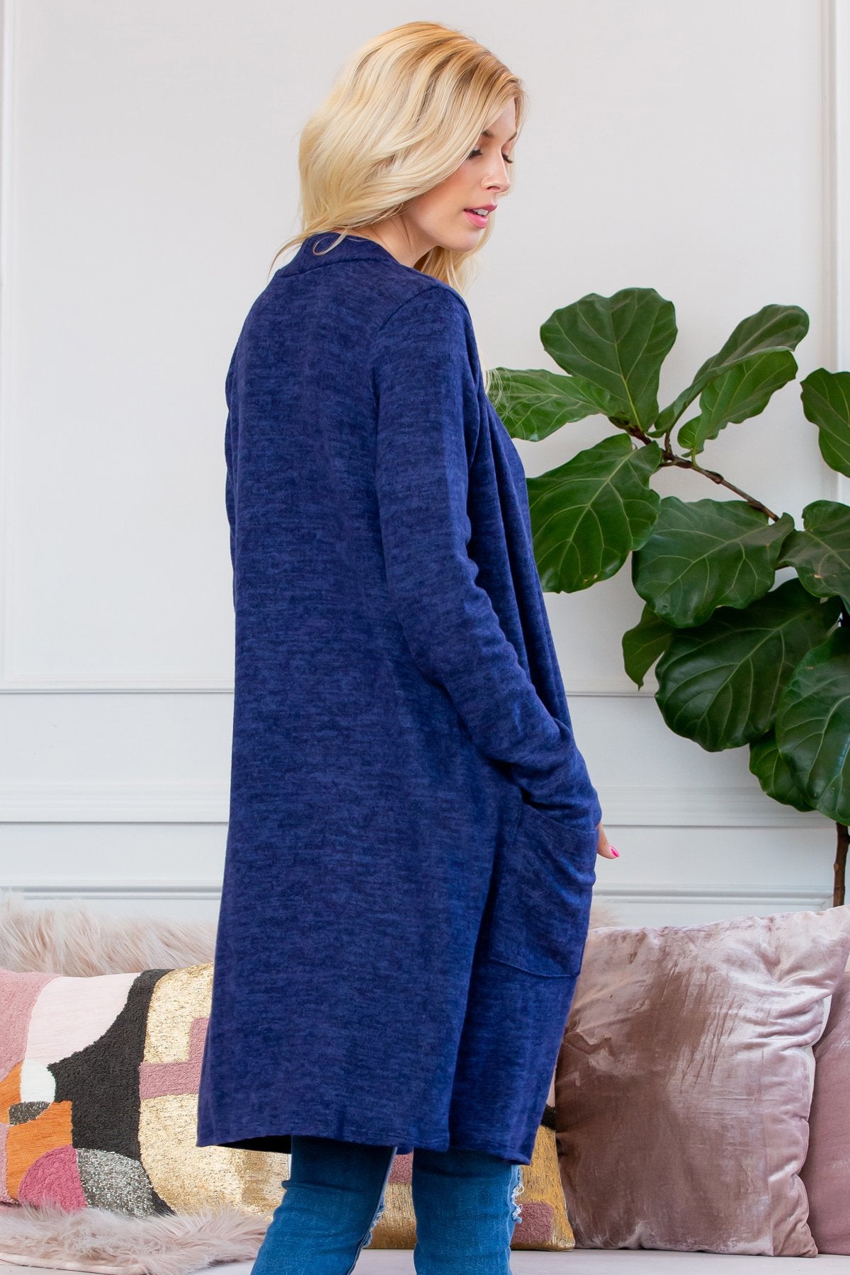 A person stands indoors near a plant, wearing Riah Fashions Brushed Sweater Pocket Cardigan in black over a white top and jeans.