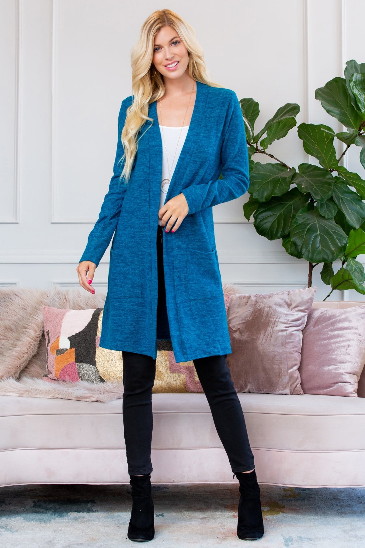 A person stands indoors near a plant, wearing Riah Fashions Brushed Sweater Pocket Cardigan in black over a white top and jeans.