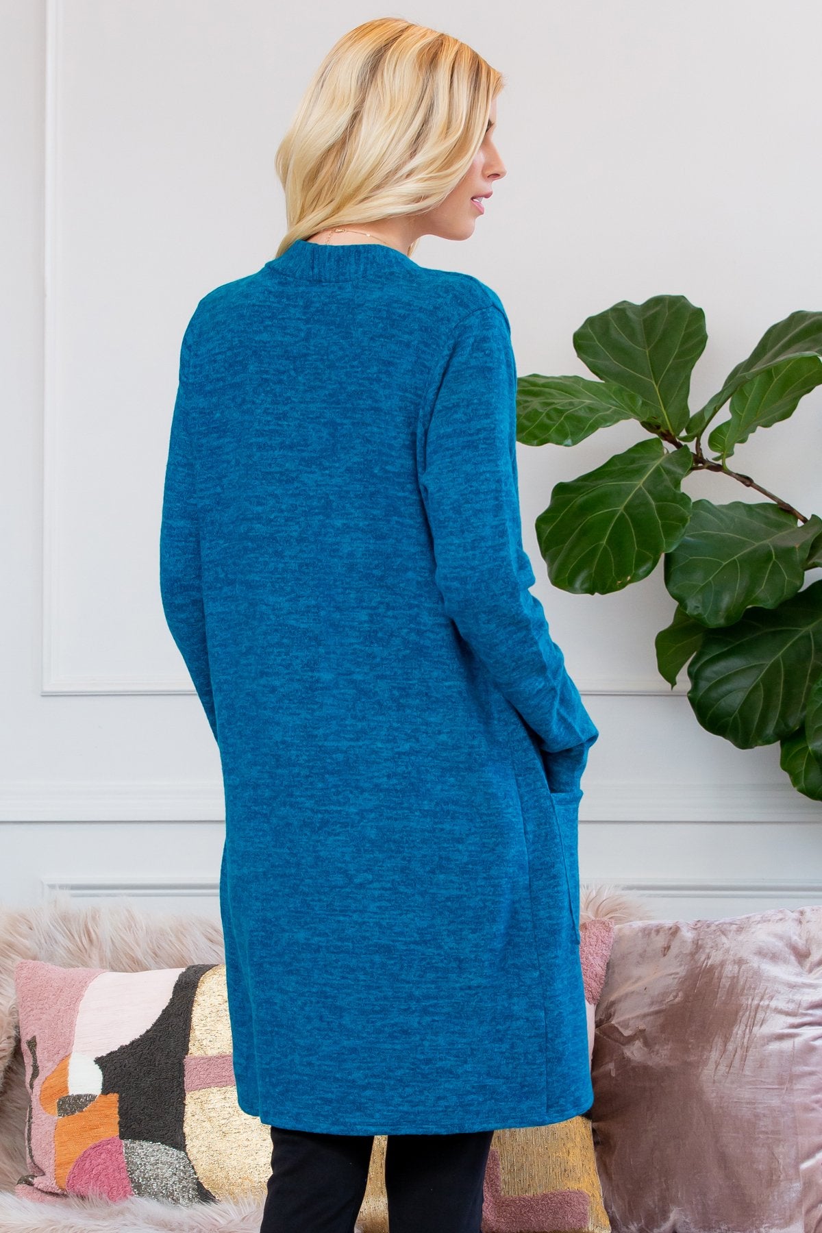 A person stands indoors near a plant, wearing Riah Fashions Brushed Sweater Pocket Cardigan in black over a white top and jeans.