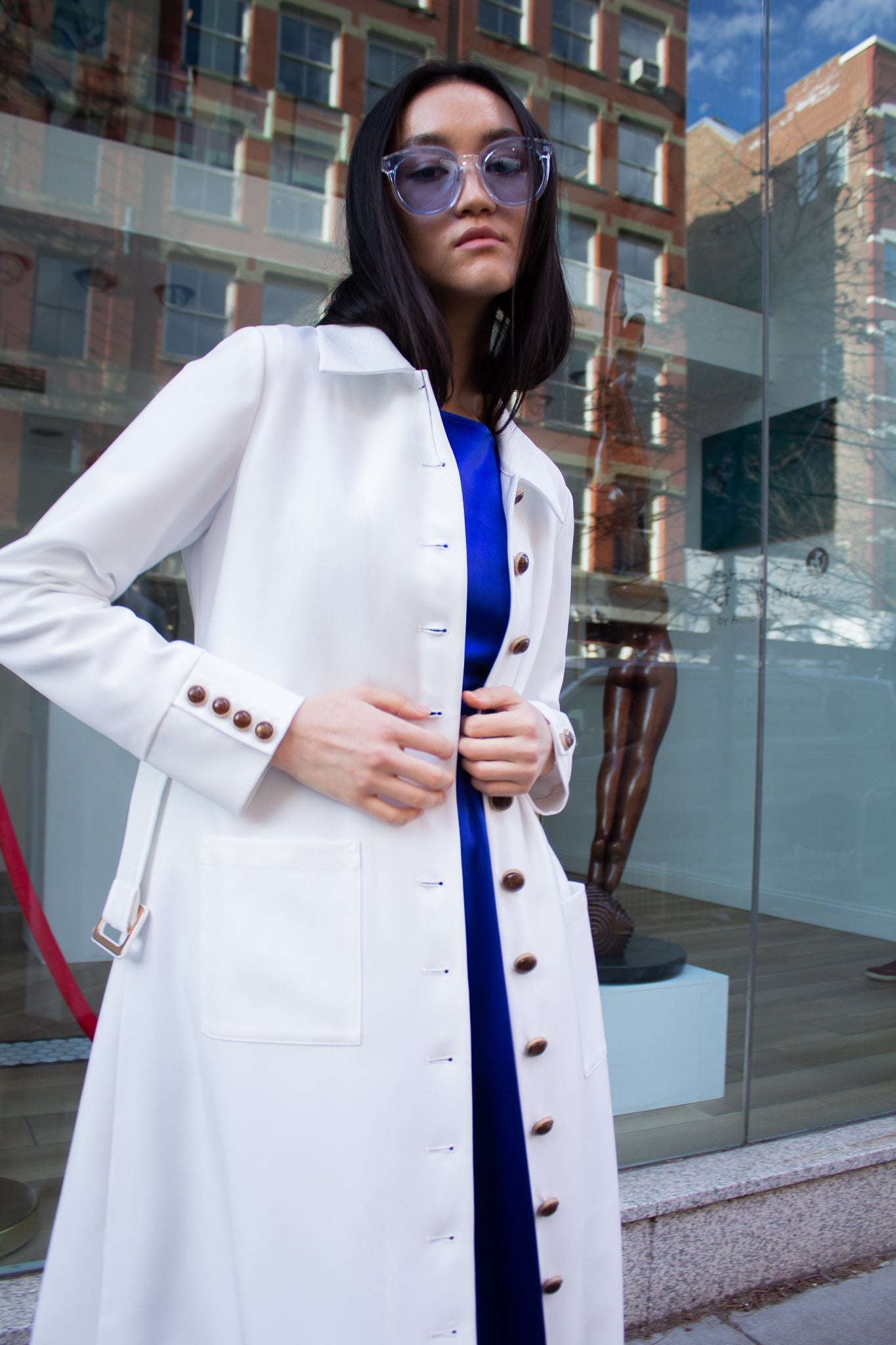 The Monique White Coat by Le Réussi® is a long-sleeve trench coat featuring a collar, button closure, matching belt, and cuff buttons.