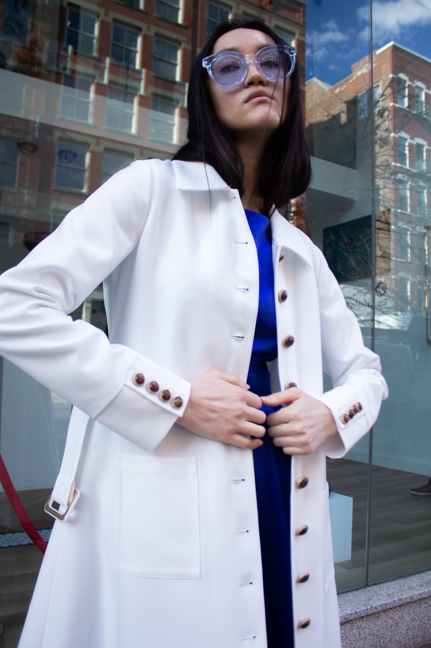 The Monique White Coat by Le Réussi® is a long-sleeve trench coat featuring a collar, button closure, matching belt, and cuff buttons.