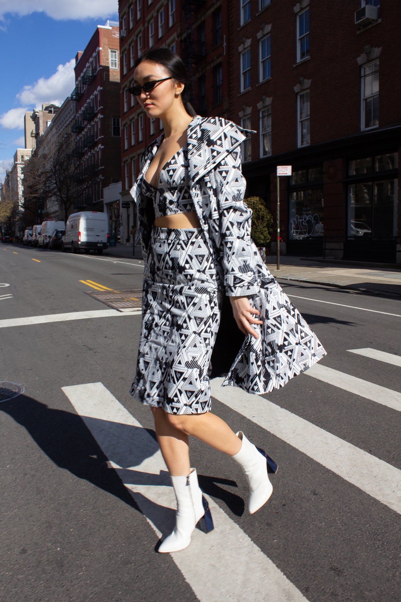 A person wearing the Amour Geometric Long Jacket by Le Réussi® and white boots stands on a sunlit sidewalk with a dark building in the background.