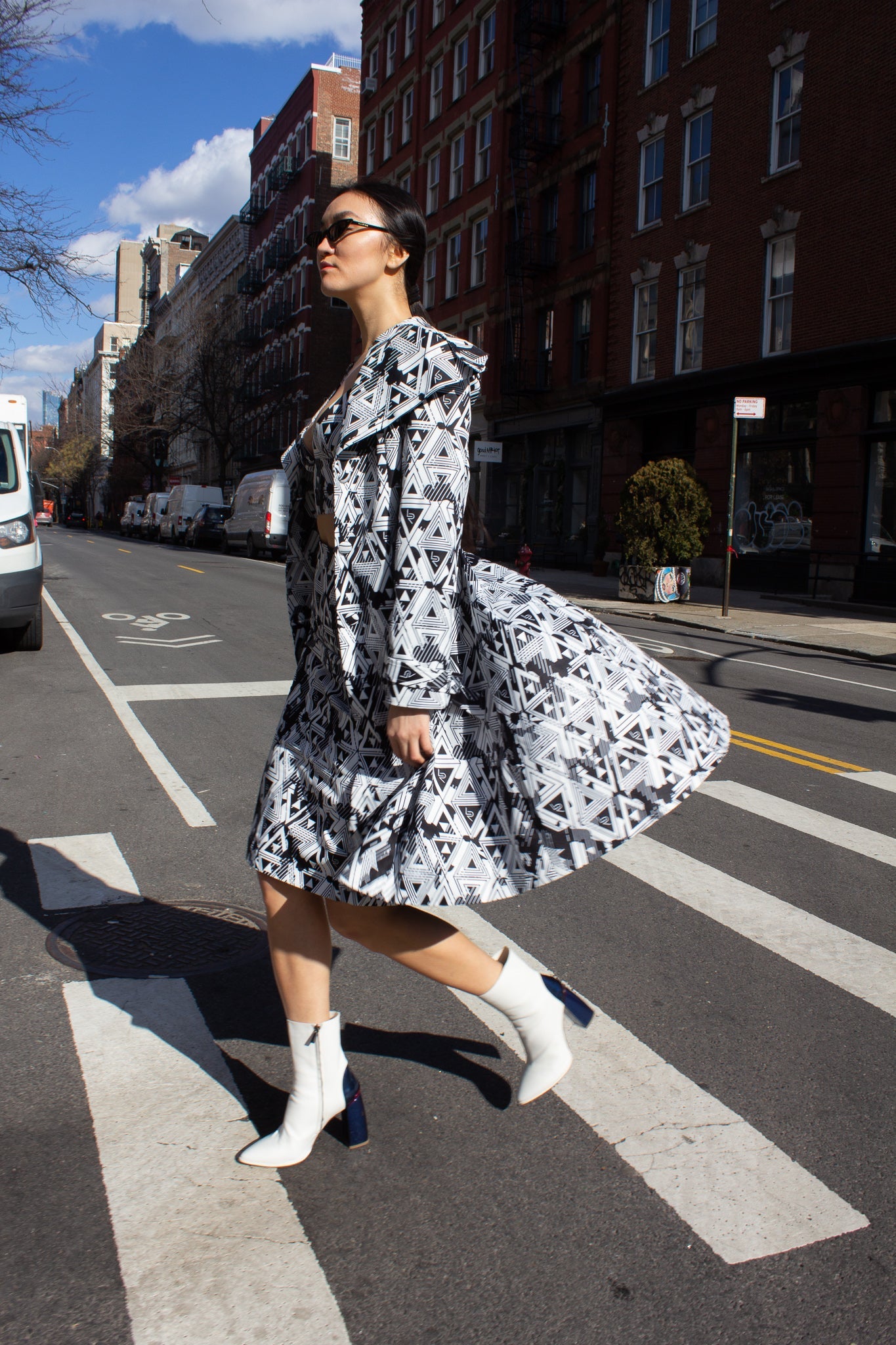 A person wearing the Amour Geometric Long Jacket by Le Réussi® and white boots stands on a sunlit sidewalk with a dark building in the background.