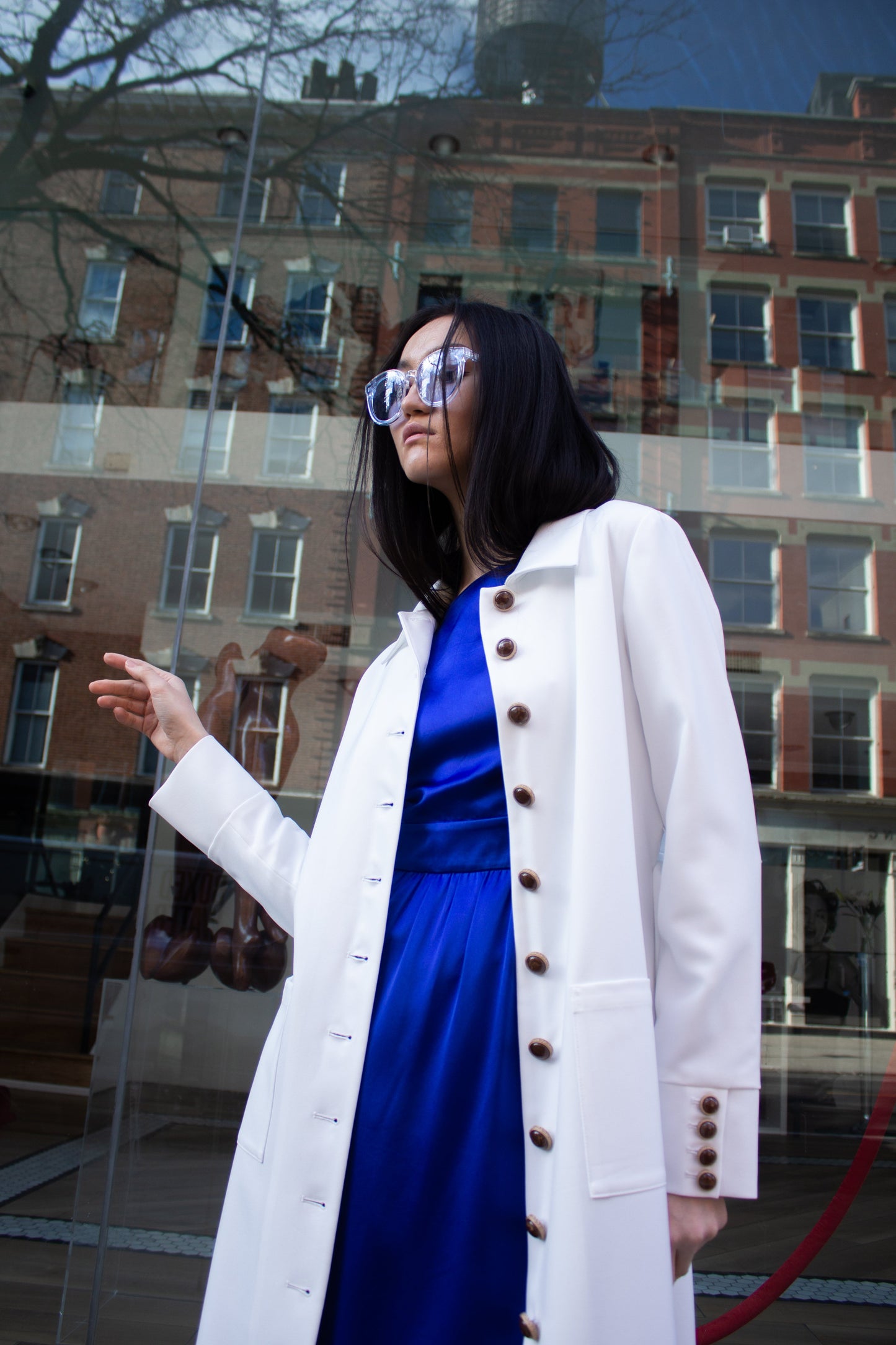 The Monique White Coat by Le Réussi® is a long-sleeve trench coat featuring a collar, button closure, matching belt, and cuff buttons.