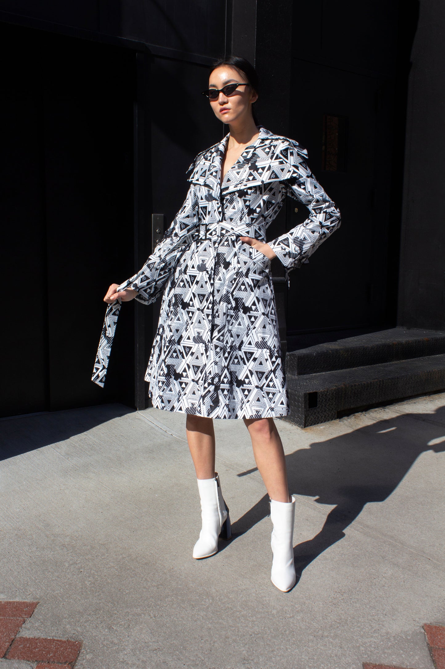 A person wearing the Amour Geometric Long Jacket by Le Réussi® and white boots stands on a sunlit sidewalk with a dark building in the background.