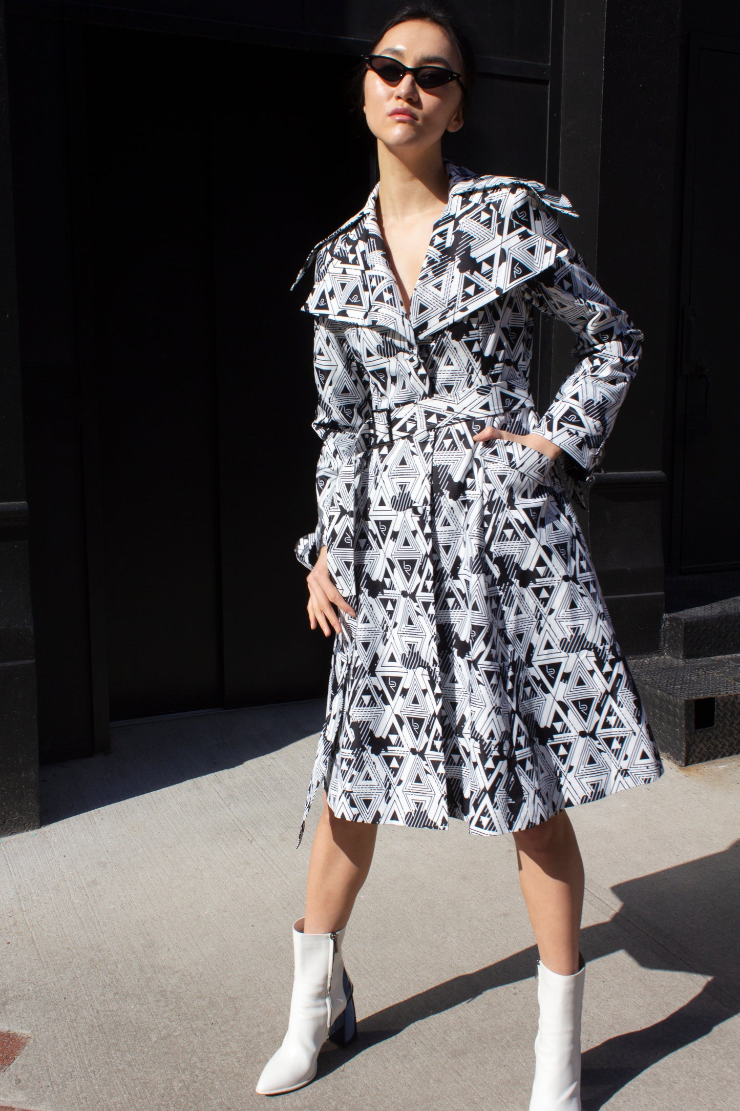 A person wearing the Amour Geometric Long Jacket by Le Réussi® and white boots stands on a sunlit sidewalk with a dark building in the background.