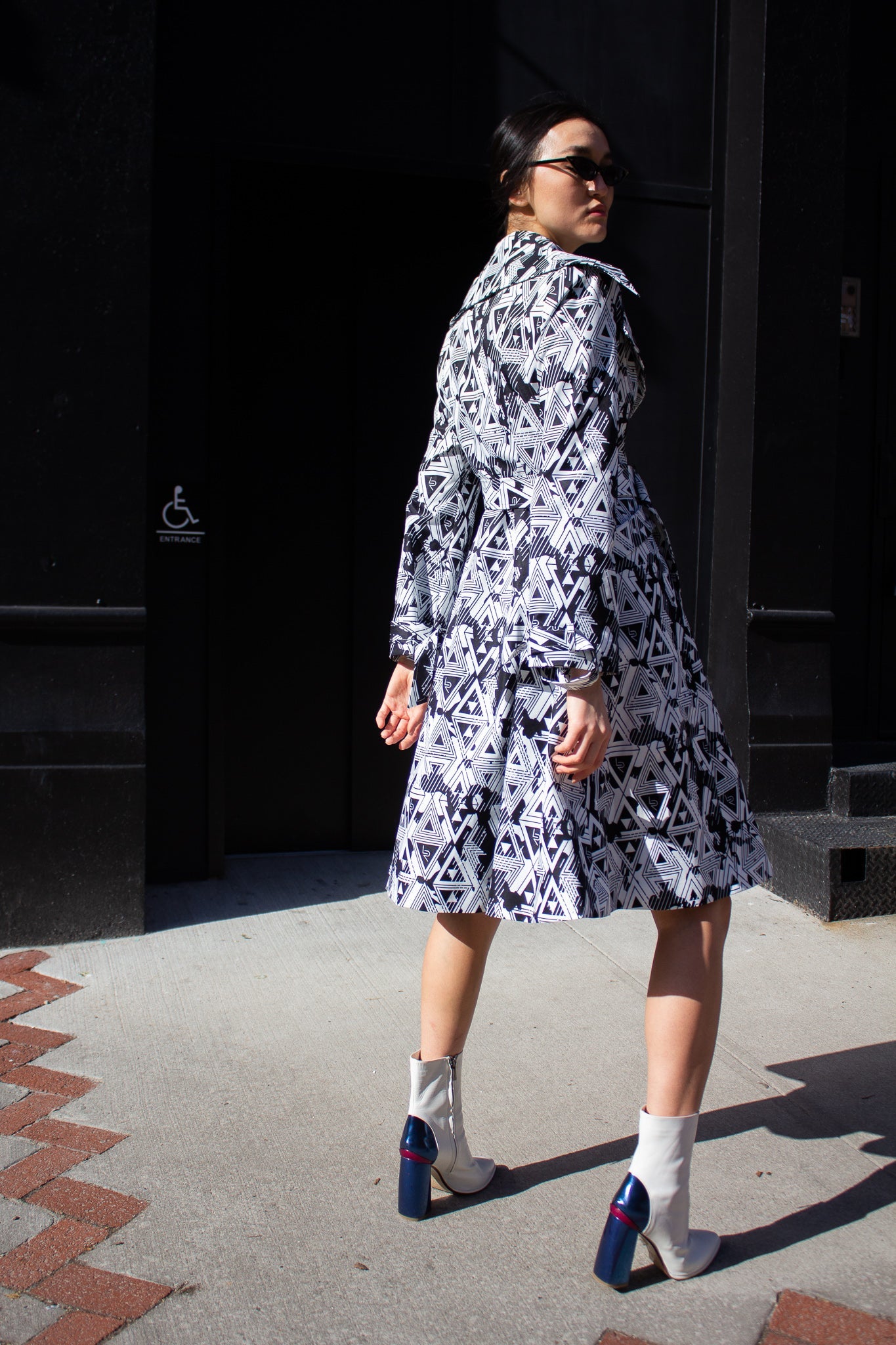 The Amour Geometric Long Jacket by Le Réussi® features a black and white geometric pattern, a belt, and a wide collar, elegantly displayed on a stand.