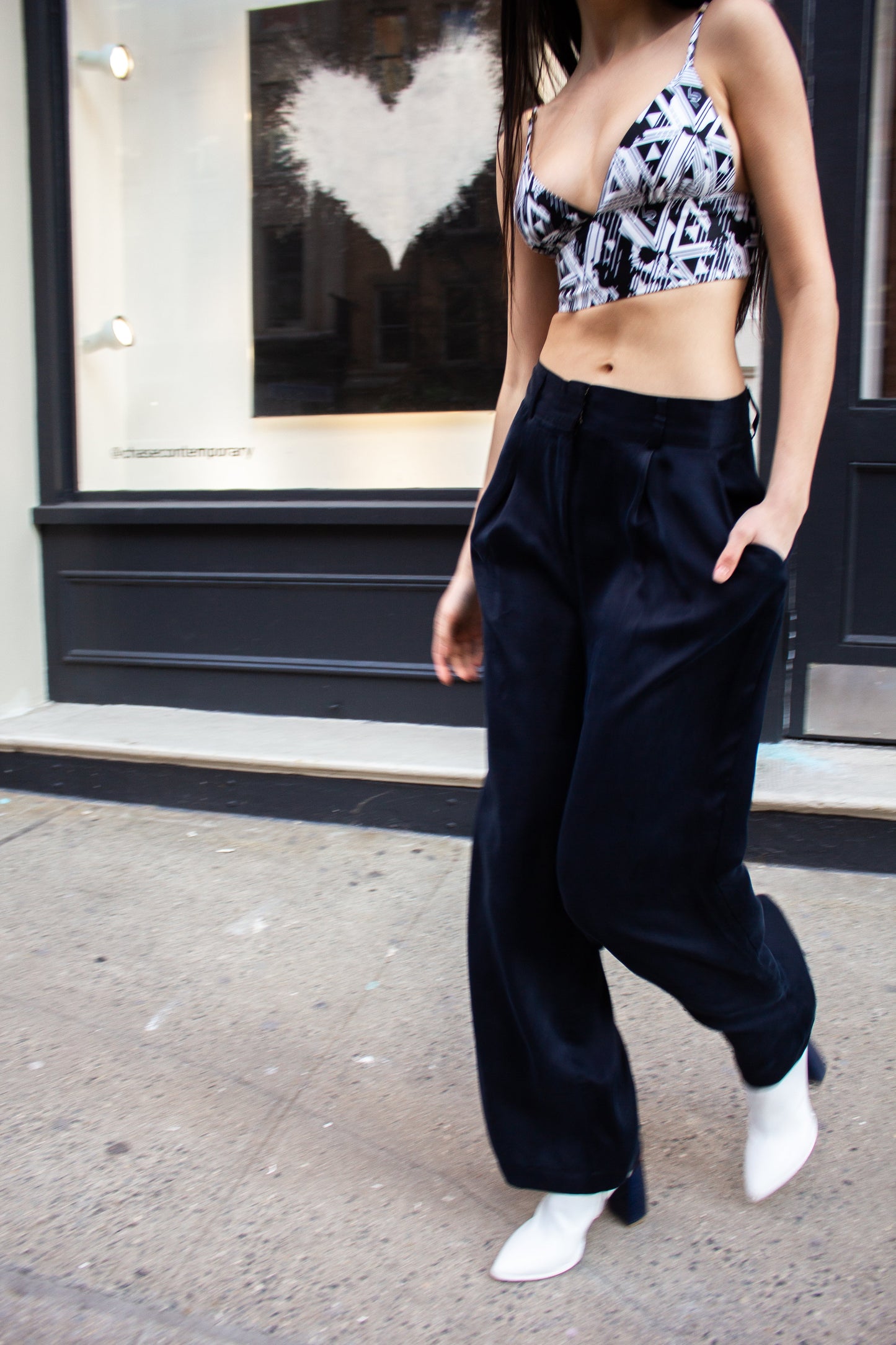 A woman in sunglasses strolls down a city street, wearing a patterned crop top, Remi Navy Pants by Le Réussi®, and white shoes, with her hands tucked in her pockets.