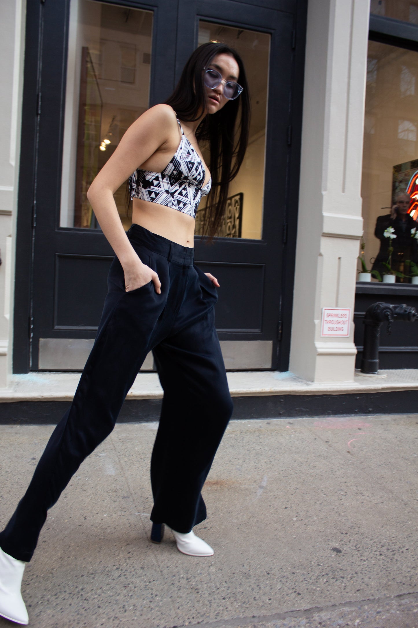 A woman in sunglasses strolls down a city street, wearing a patterned crop top, Remi Navy Pants by Le Réussi®, and white shoes, with her hands tucked in her pockets.